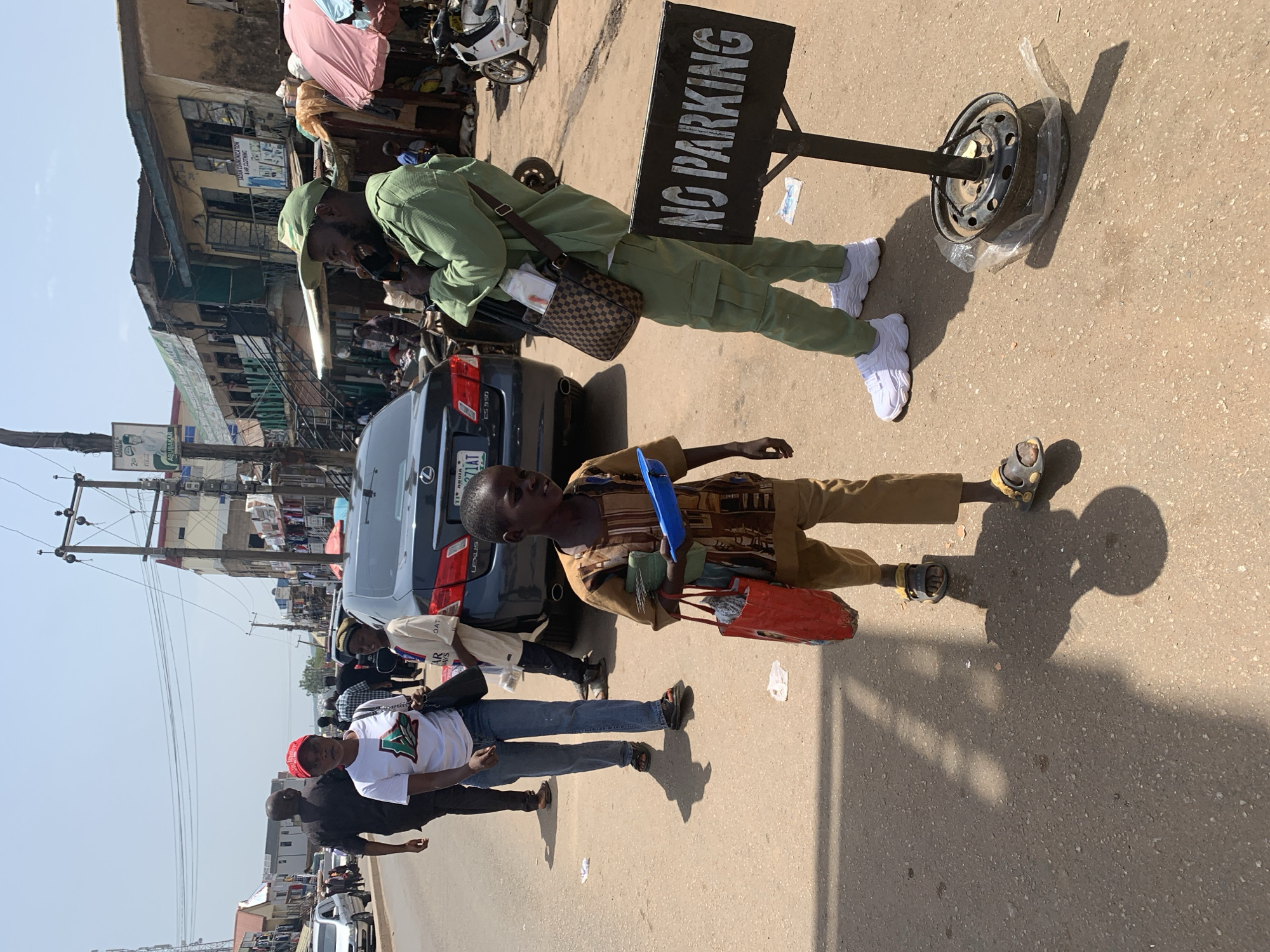 In Jos, Nigeria, a street scene unfolds with a group of individuals near a busy roadway. In the foreground, a young boy in a brown and gold patterned tunic and tan trousers walks forward, carrying a blue plastic tray and a red shopping bag. To his right, a man in a light green uniform and cap talks on his phone, a checkered brown and black shoulder bag slung across his chest. He stands near a "NO PARKING" sign. Behind the boy, a dark-colored Lexus sedan is parked. Further back, a woman in a white t-shirt and blue jeans walks away from the camera, her red cap visible. Other figures and a motorcycle are also present in the background, with shops lining the street. The scene is under a bright, clear sky.
