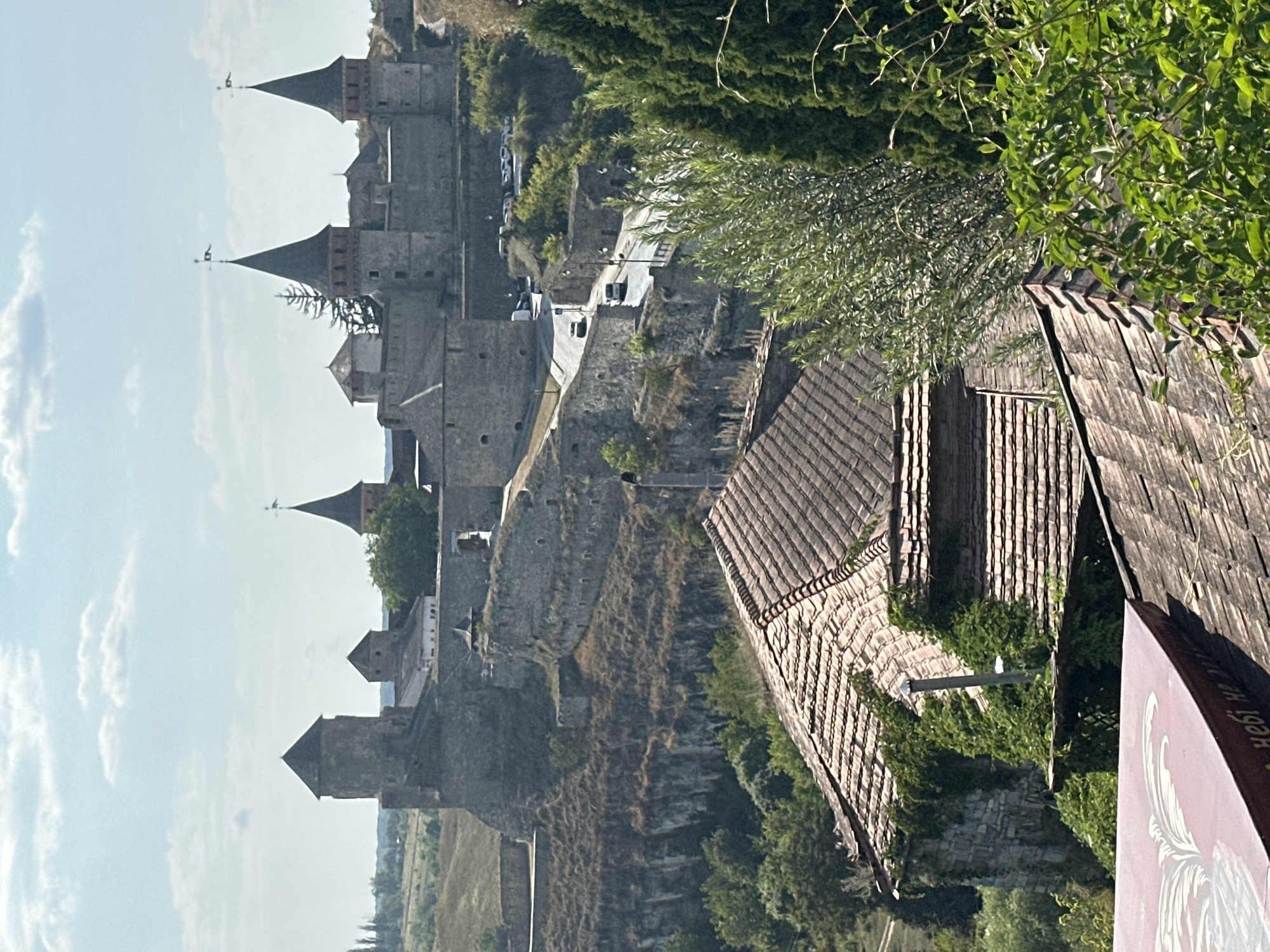 The image shows a panoramic view of the Kamianets-Podilskyi Castle in Ukraine, a historic fortress situated on a rocky promontory. In the foreground, the terracotta tiled roofs of several older buildings, partially overgrown with green foliage, dominate the lower portion of the frame. A narrow road with several vehicles, including cars and a van, winds its way up towards the castle complex, bordered by stone walls and lush green trees. The castle itself, constructed from grey stone, features multiple towers with conical roofs and flags atop them. The surrounding landscape is characterized by steep, rocky slopes covered in dry vegetation and scattered trees, indicative of a rugged terrain. The sky above is partly cloudy, with patches of blue sky visible. The scene depicts a sunny day, with ample natural light illuminating the ancient architecture and the natural environment.