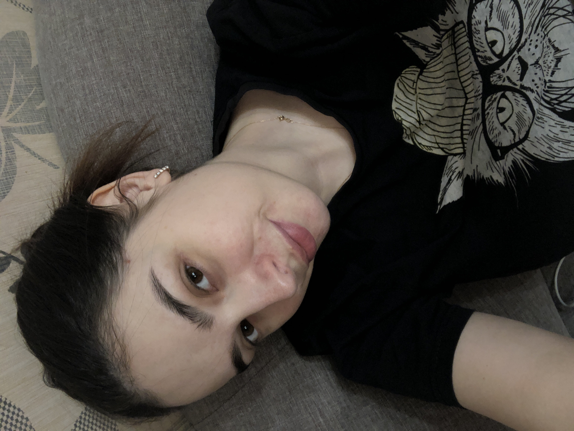A woman is lying down on a patterned gray surface. She is wearing a black t-shirt with a drawing of a cat with glasses on it. She has dark hair and is wearing a small hoop earring in her left ear and a thin gold necklace. Her head is angled towards the camera, and she is looking at the camera with a slight smile. The location is unspecified and cannot be determined from the image.