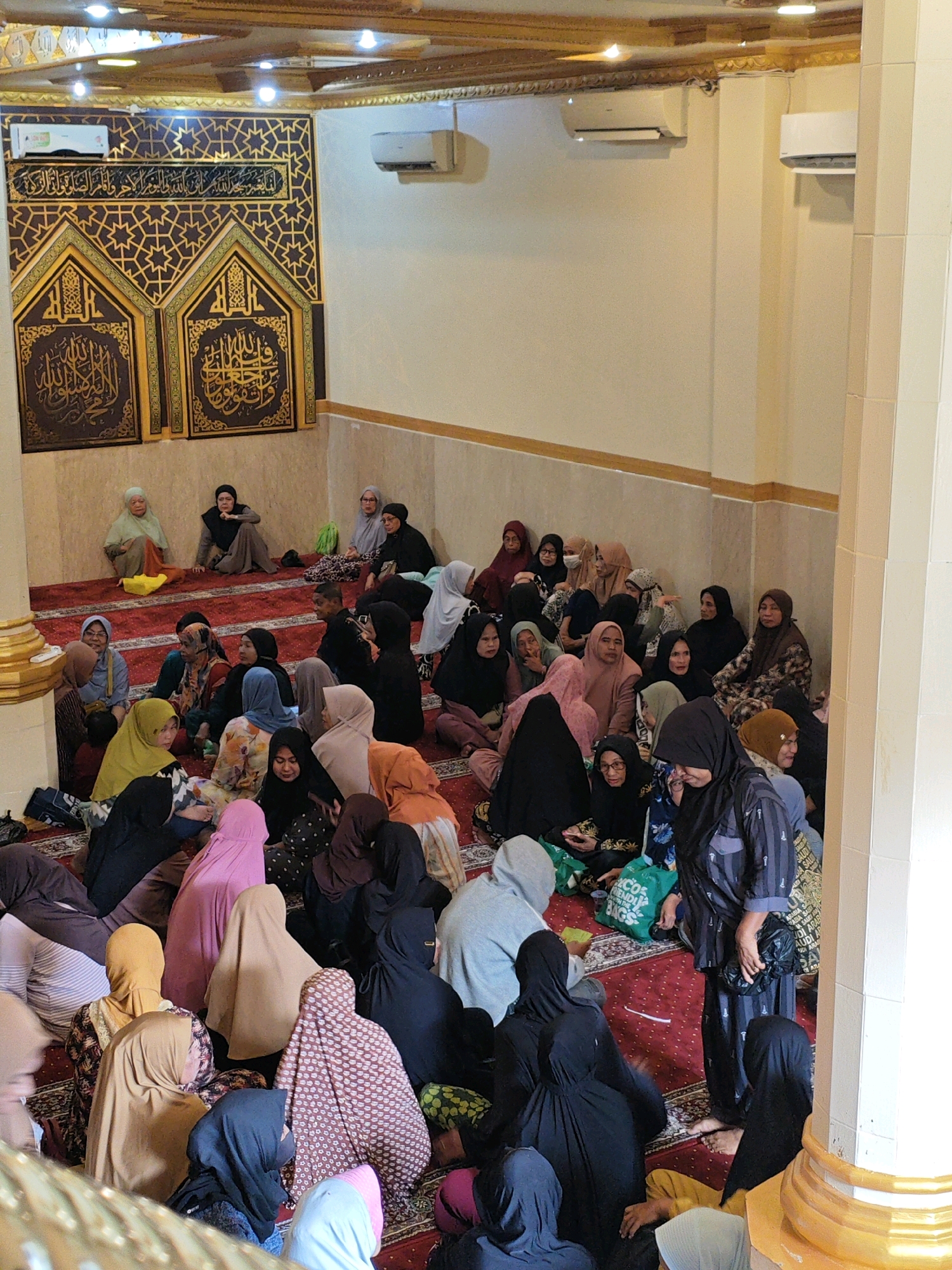 A group of approximately 30-40 women, all wearing hijabs or headscarves, are gathered indoors, seated on a red carpeted floor. They are in a room with cream-colored walls and a decorative wooden ceiling. Arabic calligraphy and geometric patterns adorn the walls above the seated individuals, suggesting the location is a religious or cultural space, likely a mosque. Two air conditioning units are visible on the upper walls. The women are of various ages and are engaged in conversation or listening, with some holding bags. The overall atmosphere appears to be calm and communal.