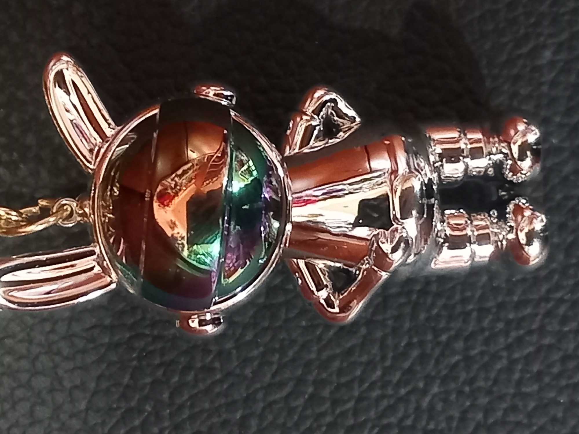 A close-up, eye-level shot shows a silver, cartoonish rabbit figurine with large ears and a reflective, iridescent face, standing on a dark, textured surface. The figurine has a gold chain attached to its head. The face of the figurine reflects a colorful, distorted image, possibly of a room or outdoor scene. The figurine is positioned in the center of the frame, with its hands on its hips. The background is out of focus, providing no discernible details about the environment or location.