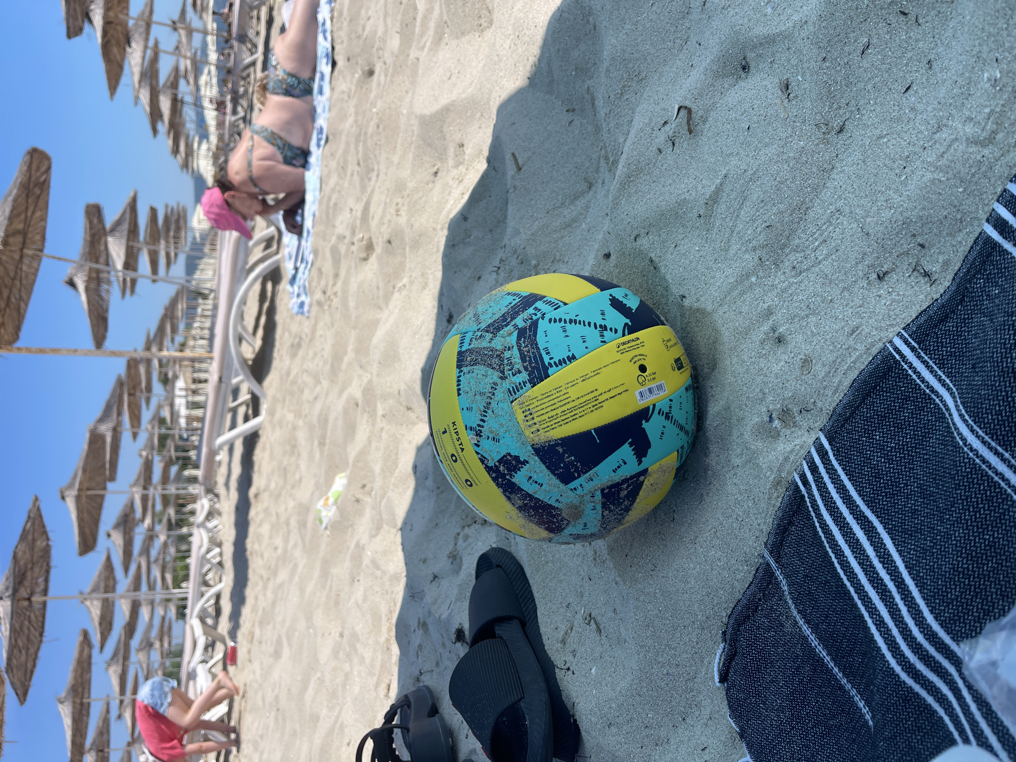 At a sandy beach in Kranevo, Bulgaria, under a clear blue sky, a yellow and blue volleyball lies on the sand in the foreground. Nearby, a black pair of flip-flops is visible. In the background, rows of beach umbrellas and lounge chairs are set up. A person wearing a pink hat and a patterned bikini lies on a towel on the sand, facing away from the camera. Further back, a child in a red shirt and denim shorts is crouched down. The overall scene depicts a sunny day at the beach with recreational activities.