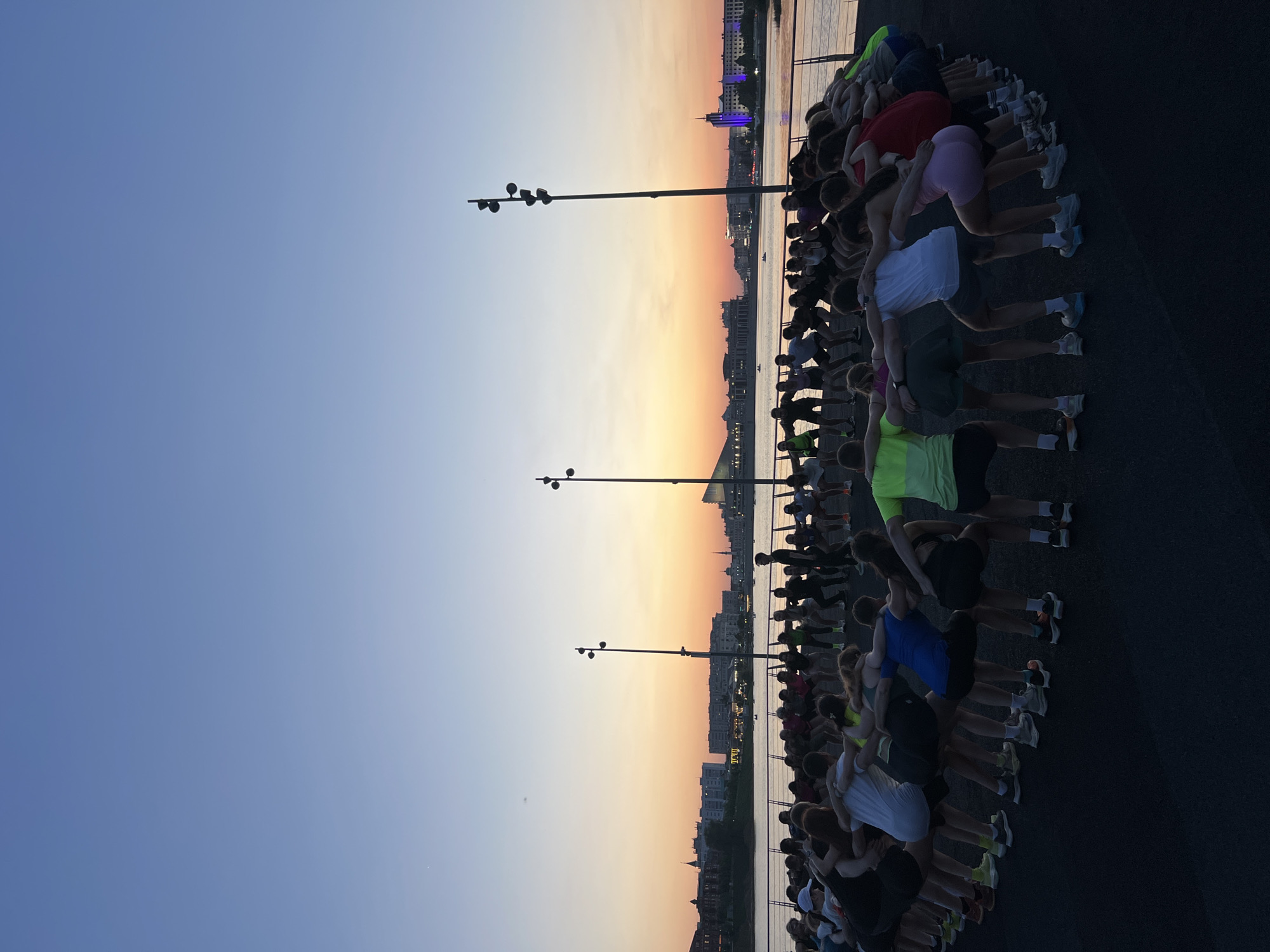 A wide-angle shot of numerous people lined up, facing the water, likely the Kazanka River in Kazan, Russia, as suggested by the city buildings visible in the background during sunset. The people are standing on a paved surface, and are touching or hugging each other. Several lamp posts are spaced out along the line of people. The sky transitions from blue to orange near the horizon.