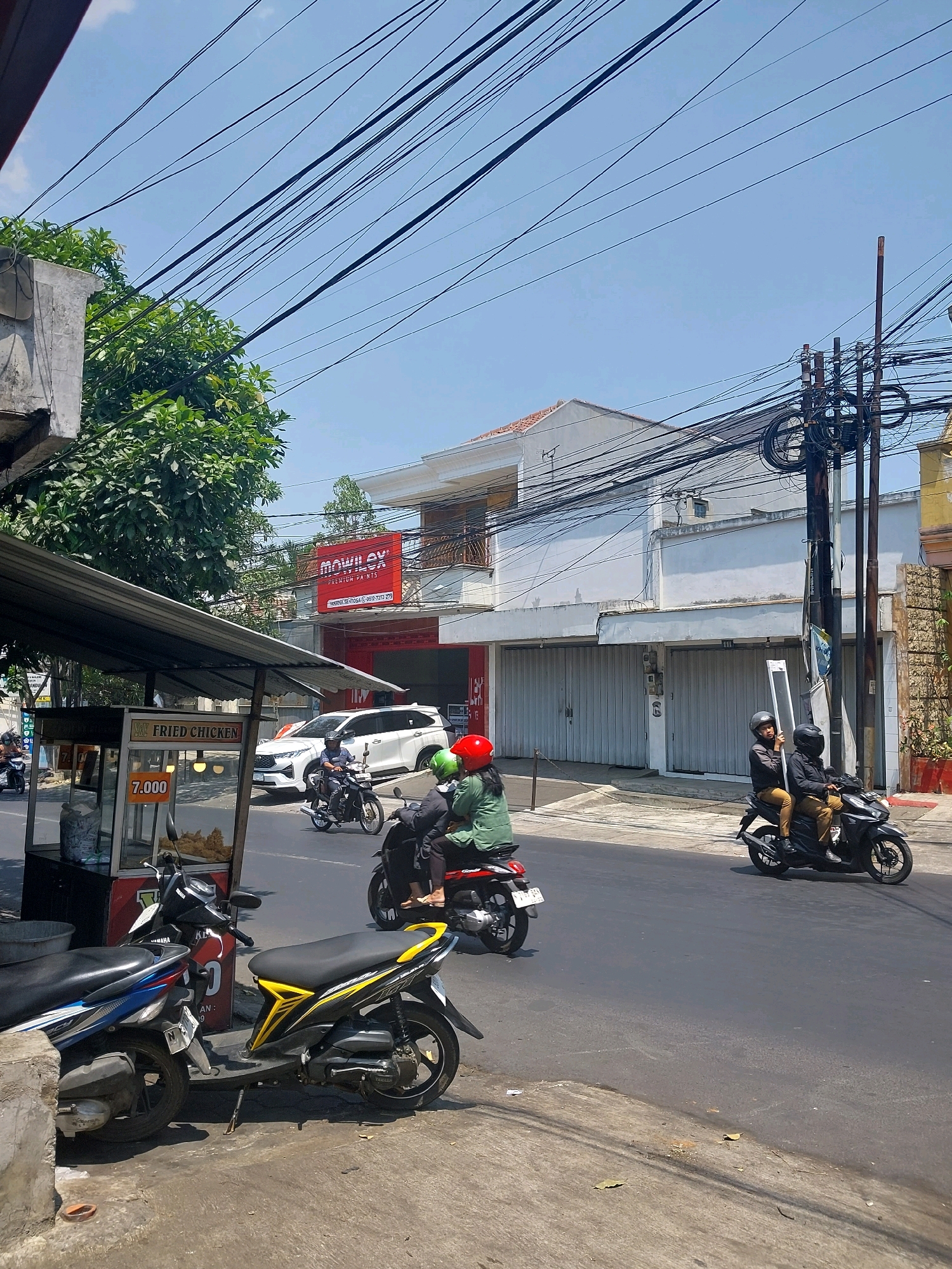 This image captures a street scene in Malang, Indonesia, on a sunny day. The foreground is dominated by two parked motorcycles, one blue and white and another black and yellow. To the left, a food stall with a sign reading "FRIED CHICKEN" and a price of "7.000" is visible under a metal awning. Across the street, a white SUV is parked.
Further down the street, several people are riding motorcycles. One motorcycle has two people wearing helmets, one red and one green. Another motorcycle carries two individuals also wearing helmets, with the passenger holding a long, flat object.
The background features a building with a prominent red sign displaying "MOWILEXX PREMIUM PANELS." Overhead, a dense network of electrical wires crisscrosses the sky, strung between utility poles. The sky is clear and blue. The overall impression is a typical urban street in Indonesia with active traffic and commercial activity.