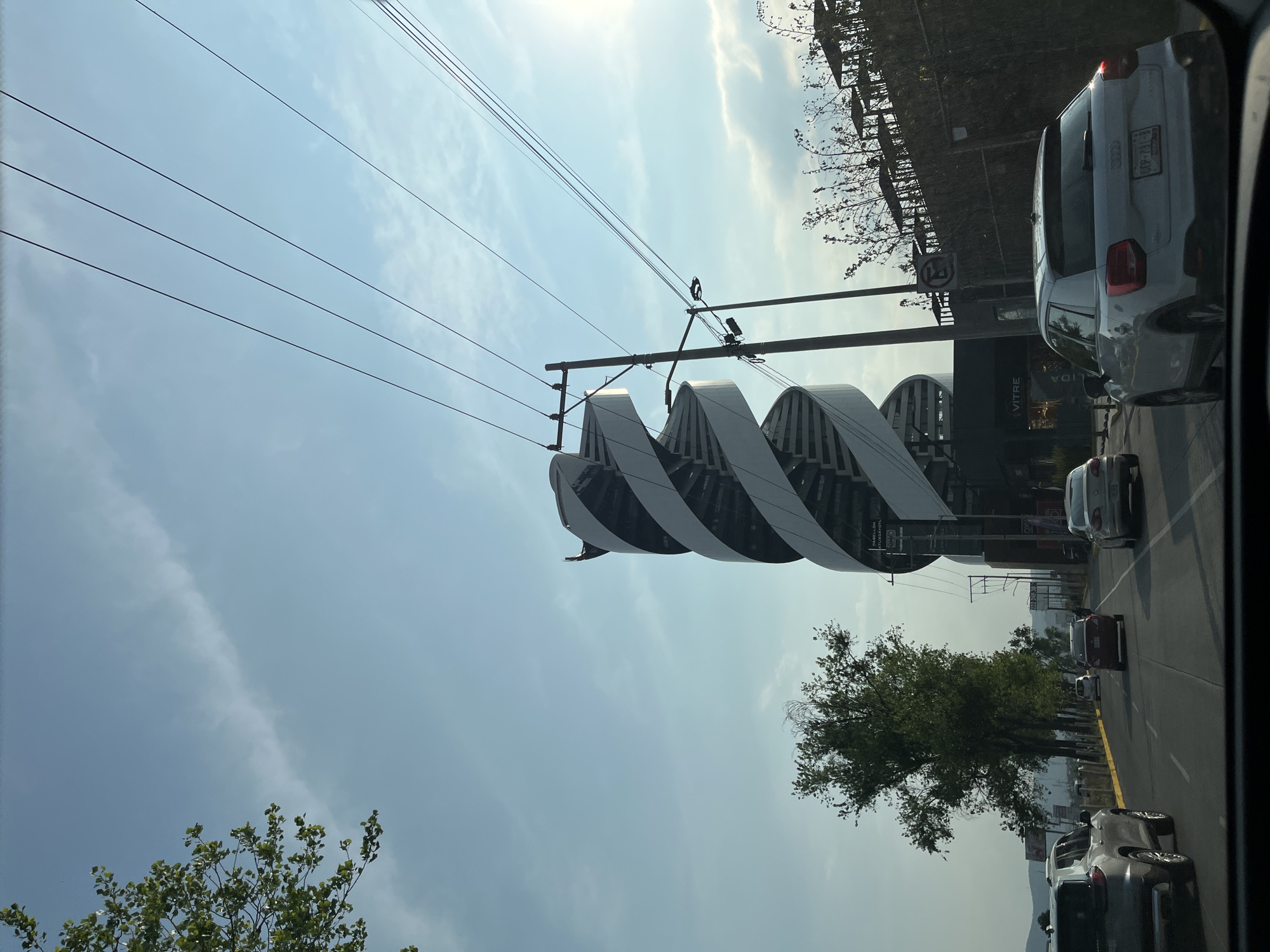 Captured from inside a vehicle, the image shows a street view in San Bernardino Tlaxcalancingo, Mexico. On the right side of the road, a unique architectural structure stands out. It consists of a series of curved, parallel fins, creating a wave-like appearance. Behind the structure are trees and glimpses of buildings, suggesting a commercial area. The road is busy with cars, including a white SUV immediately to the right, and several other vehicles further down the street. Overhead, power lines stretch across the sky. The sky is bright and clear, with thin clouds.