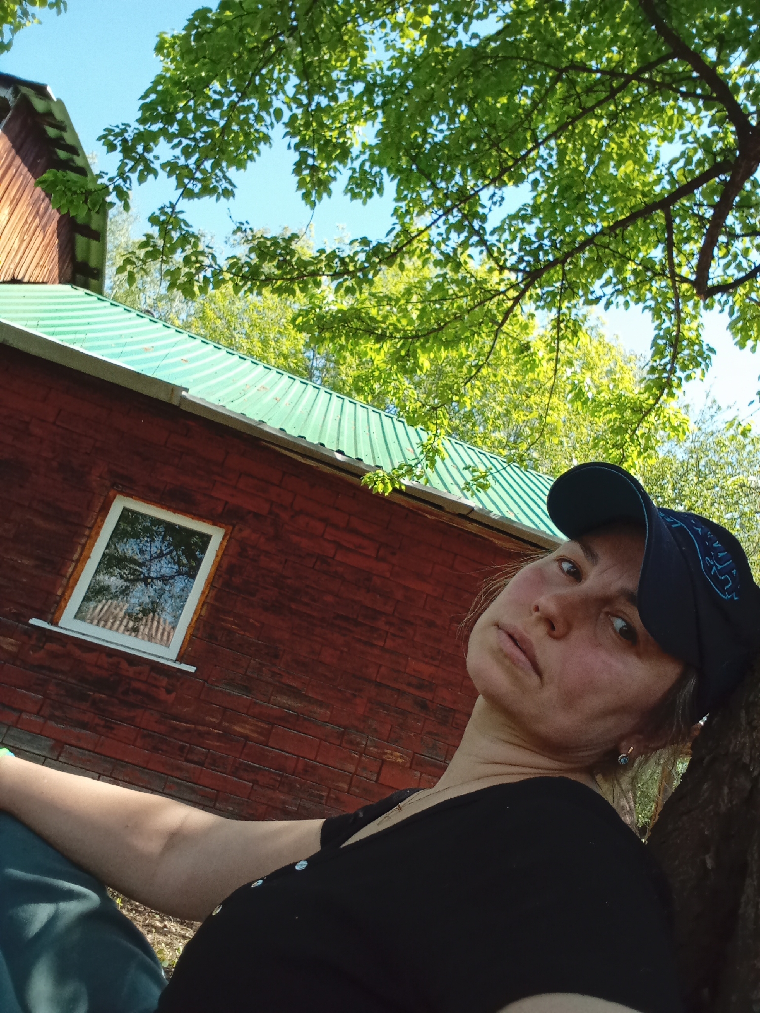 A woman in a black t-shirt and a dark blue baseball cap leans against a tree trunk. Behind her is a red wooden building with a green corrugated metal roof and a white-framed window reflecting trees. The sky is visible through the green foliage overhead. The scene appears to be outdoors during daylight. There is no verifiable evidence to confirm the location is Монастырка, Russia.