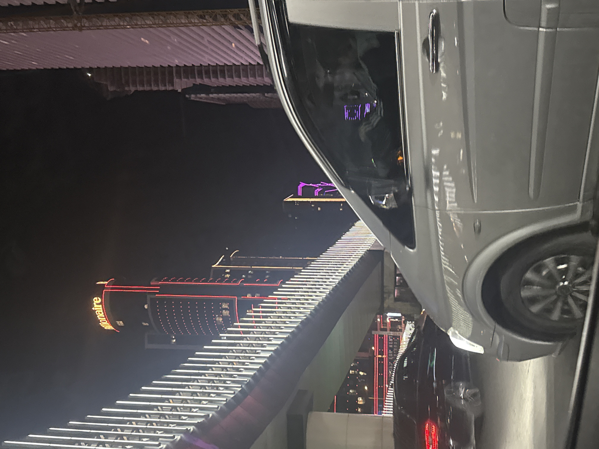 A nighttime view shows a gray van dominating the right side of the frame, with its driver-side door and front wheel visible. The backdrop features the illuminated stairs and structure leading up to the "Millionaire" casino, a prominent landmark in Batumi, Georgia. A dark-colored car is partially visible near the bottom of the frame behind the van. The lighting suggests an urban, nighttime environment.