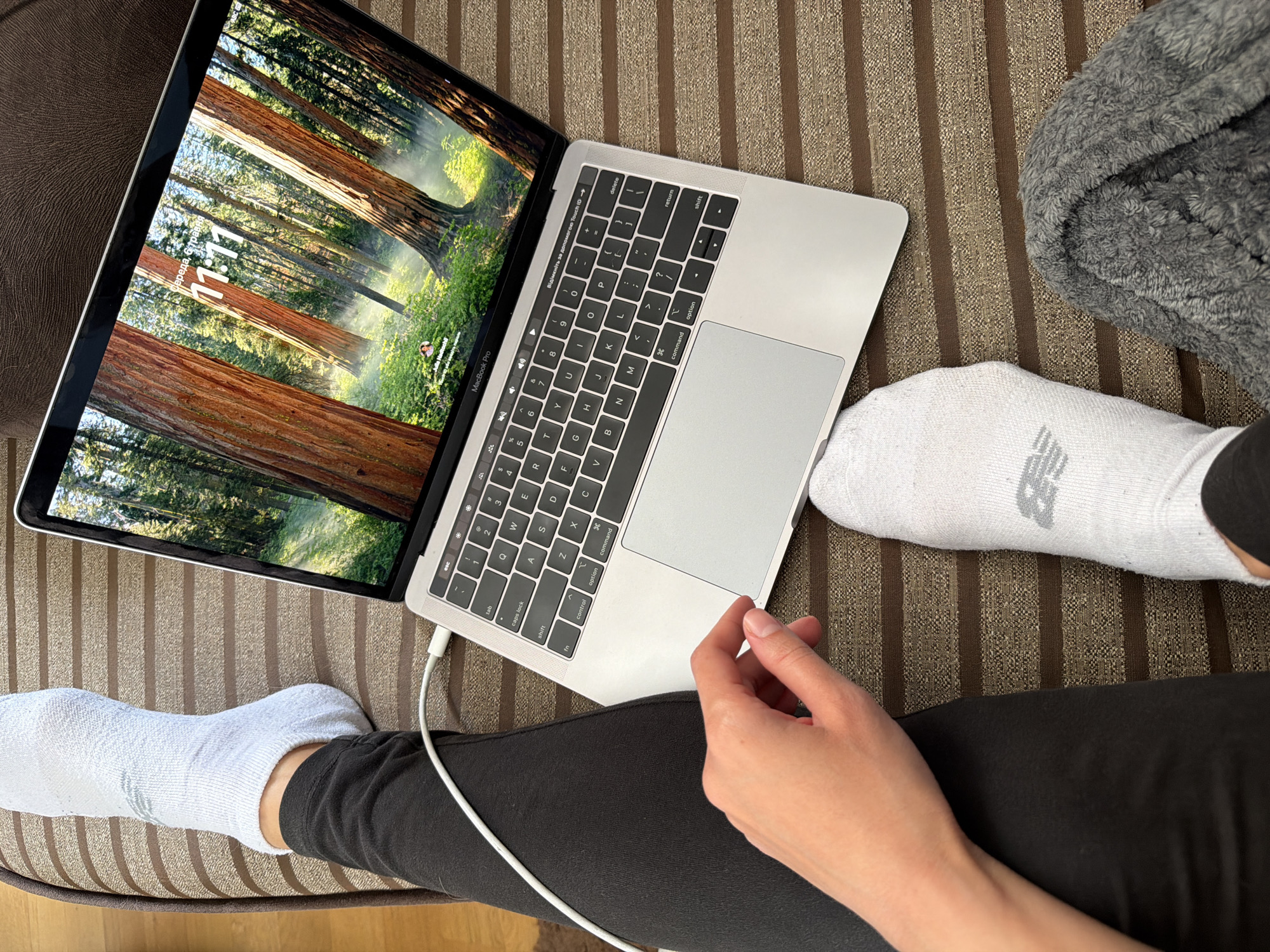 The scene depicts an individual sitting on a striped upholstered surface, possibly a couch or chair, using a MacBook Pro. The laptop screen displays a forest scene with a timestamp of "11:11" and a location possibly in Ukraine. The individual is wearing black leggings and white socks, one of which has a "New Balance" logo. One hand is visible near the laptop's trackpad. A grey blanket is visible to the right. A charging cable is plugged into the laptop. The image is a close-up shot taken from an elevated angle, likely overlooking the user in a casual, domestic setting.