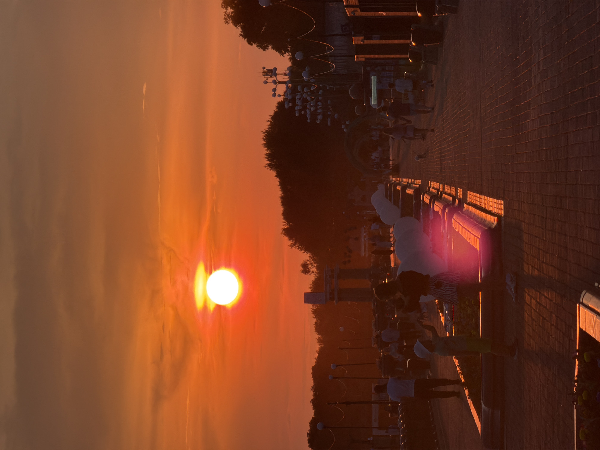 The image depicts a sunset scene in Birobidzhan, Russia, featuring a vibrant orange sky with the sun prominently positioned. In the foreground, a paved walkway with brick patterns is visible, reflecting the warm hues of the sunset. Several individuals are present, silhouetted against the light. On the left side, a woman and a child are standing near a planter bed. The woman is wearing a patterned skirt and a dark top, while the child is in a light-colored shirt and shorts, with a white hat. Further along the walkway, other people are seen walking, some in pairs, and a small dog is also visible. In the mid-ground, there is a row of decorative elements, possibly fountains or planters, with large white spherical objects. Beyond that, a line of trees forms the horizon, with a concrete structure in the distance. The overall atmosphere is one of a public promenade or park area during twilight.
