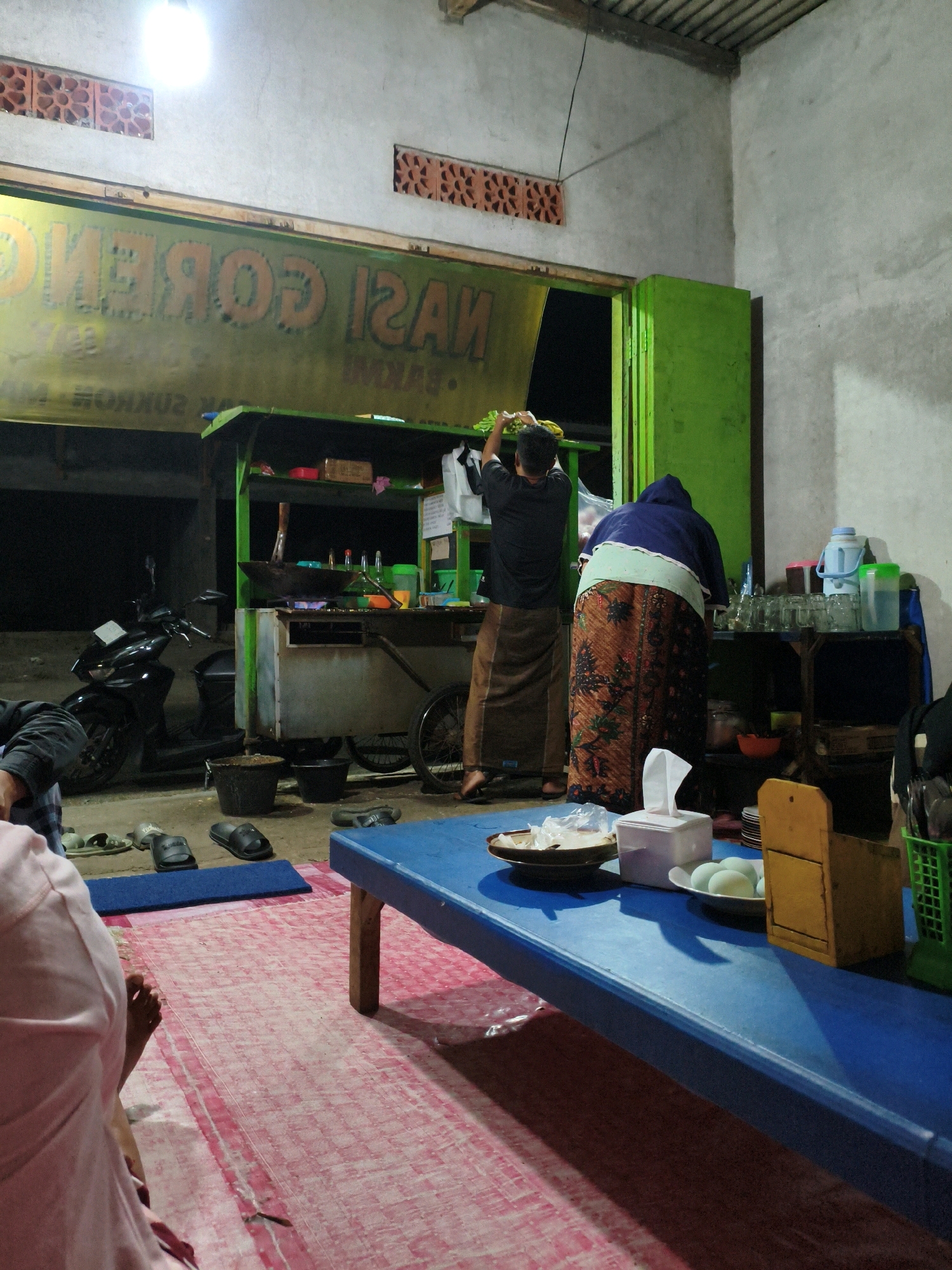 The image depicts an outdoor scene at night, likely a food stall, in Krajan Lor, Indonesia. A woman in a blue headscarf and a patterned skirt stands at a green food cart, facing away from the camera, arranging items on a shelf. A man wearing a dark shirt and a brown sarong is also at the cart, facing the cart and reaching for something. In front of the cart, a motorcycle is parked. The food cart has a large yellow sign above it with text that appears to read "NASI GORENG", indicating fried rice. A blue table is in the foreground, with a tissue box, plates, and what appear to be eggs on it. A pink mat covers the ground in front of the table. The environment suggests a casual, roadside dining setting.
