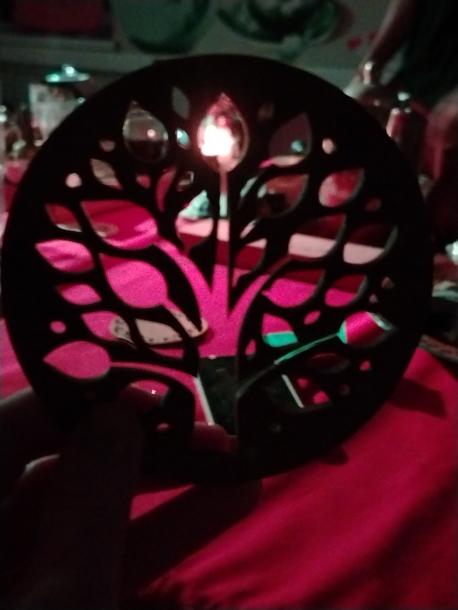 A person is holding a circular, dark-colored decorative object with a tree design cut out of it. A candle is placed behind the object, illuminating the cut-out design with a warm light. The background appears to be a room with various objects and possible people present. The scene is bathed in a reddish-pink light. There is no clear evidence to confirm that this image was taken in Chaikovsky, Russia.