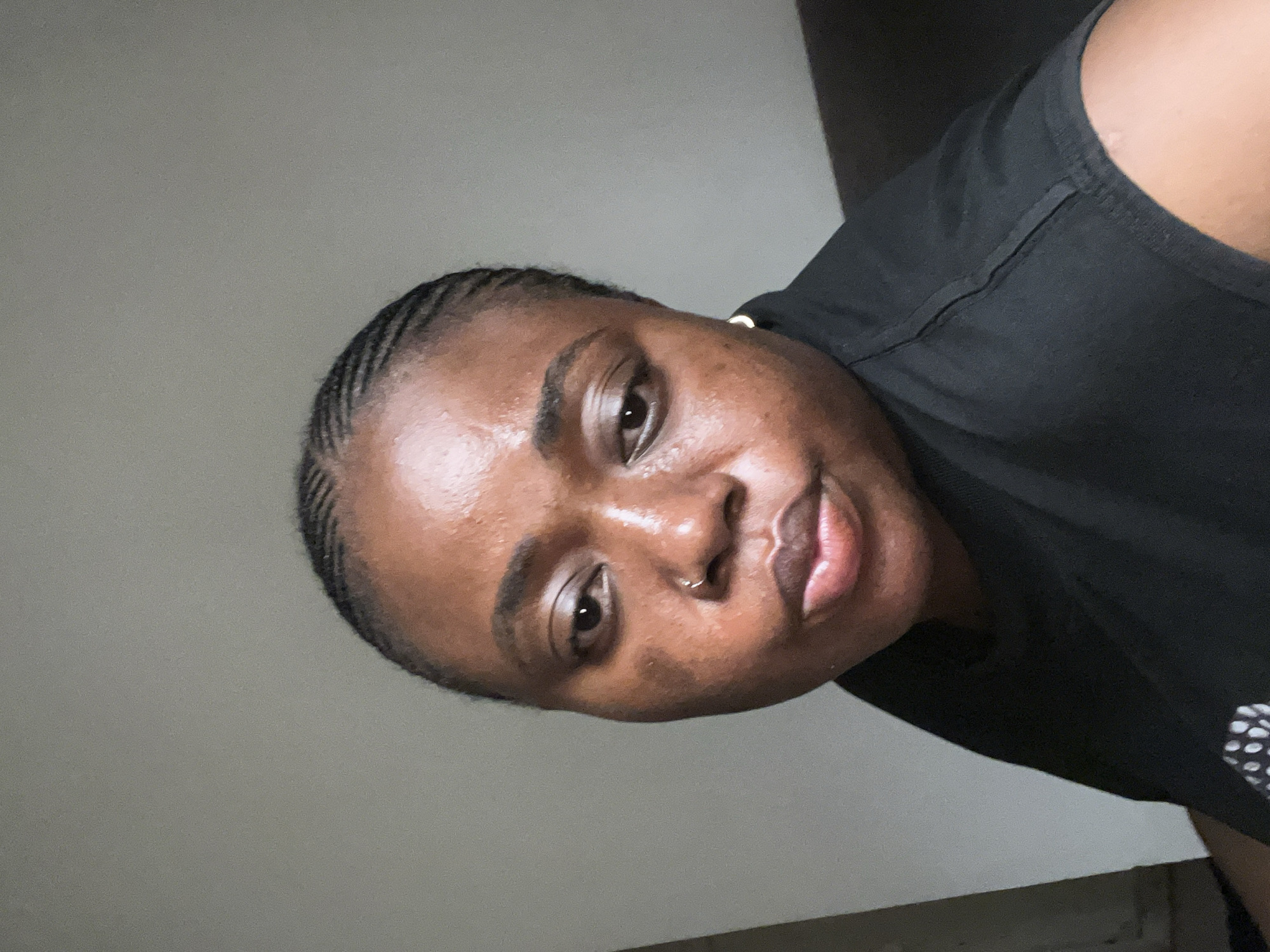 The image shows a medium shot of a Black woman in Abuja, Nigeria. She is looking directly at the camera with a neutral expression. Her hair is braided in cornrows. She is wearing a black t-shirt. The background is a plain grey wall, suggesting an indoor setting. There is a hint of furniture in the lower right corner, possibly a couch or chair. The lighting is soft and diffused, casting subtle shadows on her face.