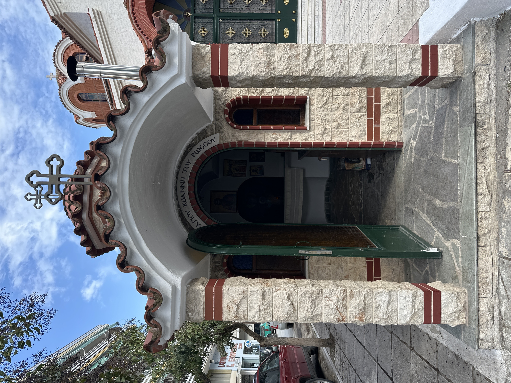 The image shows the entrance to a Greek Orthodox Church in the city of Serres, Greece. The doorway is arched with white walls, brick accents and a tiled roof. A green door is partially open leading into the church's interior. Outside the doorway are stone blocks and a sidewalk with a tree and a car parked nearby. The sky is visible in the background.