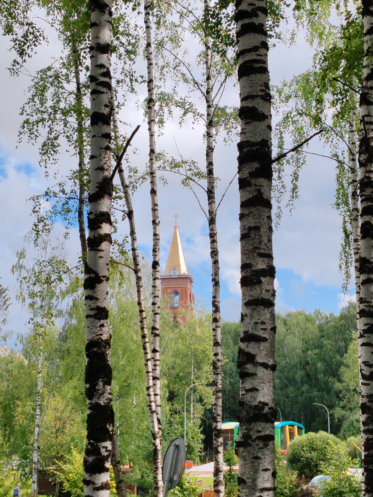 The image captures a view through a grove of birch trees, with a church tower visible in the background. The church tower is tall and slender, topped with a golden spire and a cross. It appears to be constructed of red brick and features arched windows. Surrounding the church are lush green trees, forming a dense forest. The sky above is a mix of blue with scattered white clouds. In the foreground, the distinct black and white bark of the birch trees dominates the frame, with their leaves forming a canopy. There are also glimpses of some inflatable structures, possibly a playground, to the right of the church. The overall setting suggests a park or recreational area adjacent to a religious building within the city of Syktyvkar, Russia. A single person can be seen in the lower left corner of the image, walking.