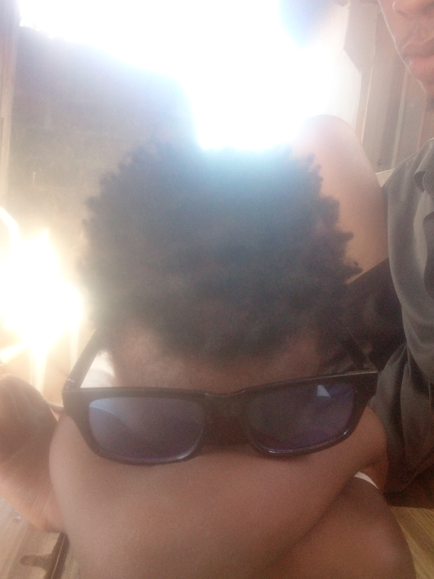 A person is seen from a close-up, low-angle perspective. The person has dark, curly hair and is wearing black sunglasses. Their skin tone is medium brown. A portion of another person is visible to the upper right, wearing a dark gray garment. The background is blurred by overexposure to light. The location is Unknown, Unknown.