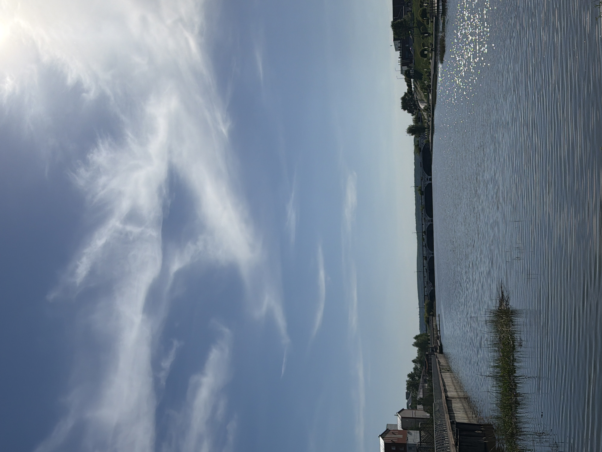 The image shows a cityscape in Votkinsk, Russia, alongside a body of water, possibly a river or lake. The sky is predominantly blue with wispy, cirrus clouds. On the right side, the water reflects sunlight, creating a shimmering effect. A shoreline with vegetation is visible along the water's edge, with docks or embankments lined along it. In the distance, a bridge can be seen spanning across the water. Buildings and trees line the far bank.