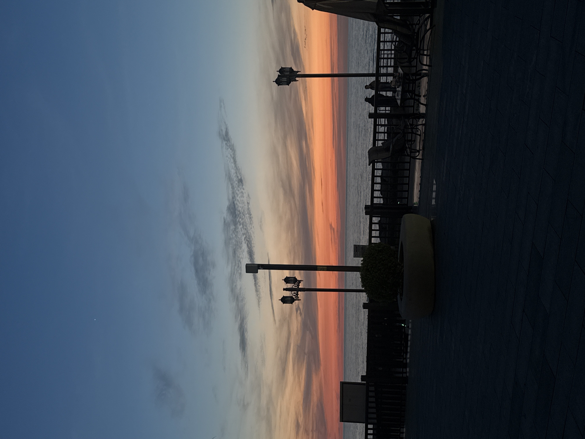 The image captures a serene twilight scene along a waterfront promenade in Batumi, Georgia. The sky dominates the left side, displaying a stunning gradient of colors indicative of either sunset or sunrise. Hues transition from a deep blue at the top left, through soft grays of scattered clouds, to vivid oranges and pinks hugging the horizon line over a calm body of water, likely the Black Sea. A small, bright white dot, possibly a star or planet, is visible in the upper blue sky.
Along the right side of the frame, a dark promenade with paved tiles runs parallel to the water. Several decorative streetlights, featuring ornate triple lanterns, stand as prominent silhouettes against the vibrant sky. People are seated at outdoor tables and chairs, also appearing as dark figures, suggesting they are dining or relaxing while enjoying the view. A dark railing or fence separates this seating area from the water. Partial views of an awning and a planter with shrubbery are also visible. The foreground elements are deeply shadowed, emphasizing the bright expanse of the sky and water. The overall atmosphere is peaceful and contemplative, with no discernible text.