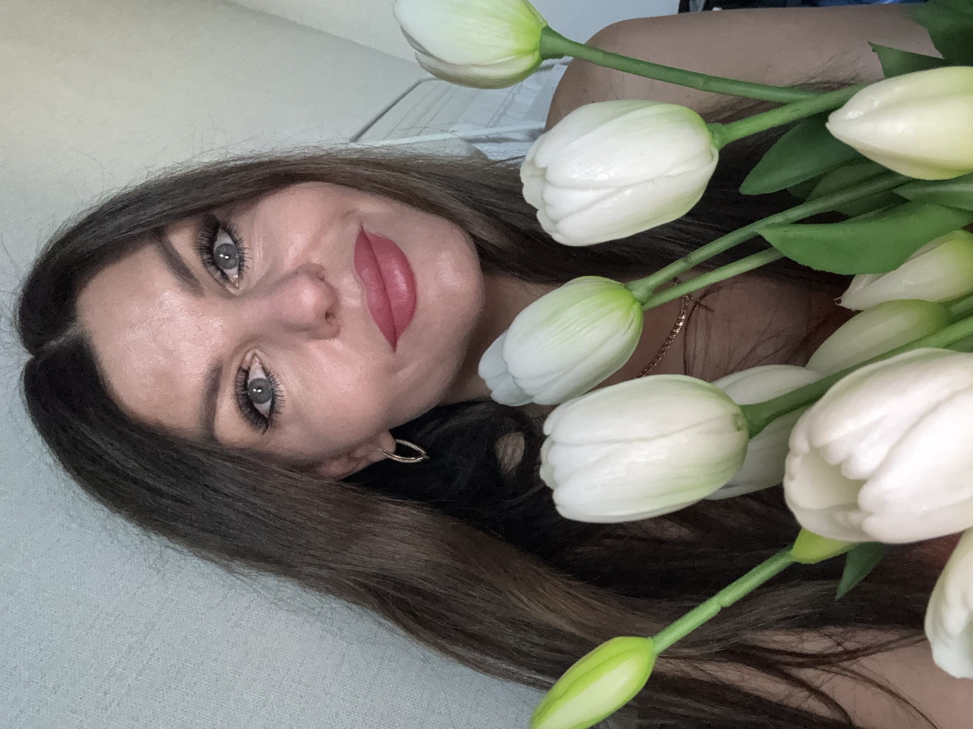 A woman with long brown hair lies on a light-colored surface next to a bouquet of white tulips. She has light skin, grey eyes, and red lipstick. She wears a gold earring and a delicate gold necklace. The background is plain and out of focus. There is no explicit indication of the location being Chaikovsky, Russia.