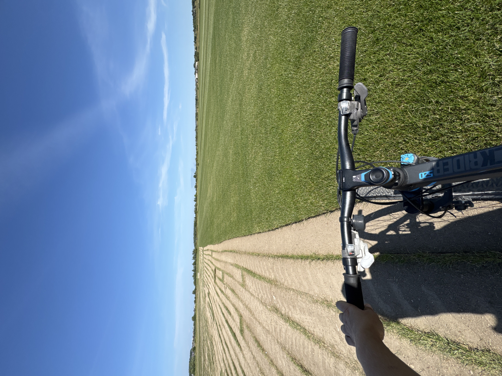 In Kempelevo, Russia, a first-person perspective shot captures a hand gripping the handlebar of a bicycle. The bicycle is positioned on a dirt path with tire tracks, adjacent to a meticulously maintained, striped green lawn. The landscape extends to the horizon, meeting a clear blue sky. In the distance, scattered buildings and trees suggest a rural or semi-rural setting.