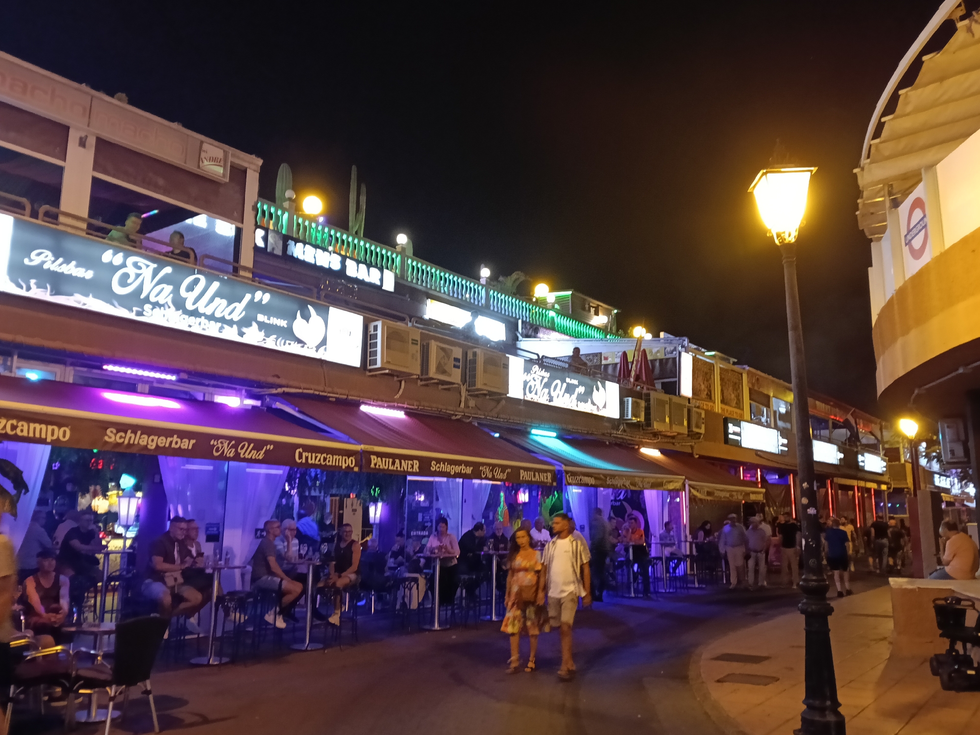 This is a nighttime street scene in what is likely San Bartolomé de Tirajana, Spain. The street is lined with bars and restaurants, all illuminated with bright neon lights. A couple walks down the street in the center of the frame, and numerous other people are seated at tables outside the establishments, engaged in conversations and drinking. The bars have signs in German. A tall lamppost is prominently placed on the right side of the frame. A London Underground logo is visible on a building corner on the right side of the frame.
