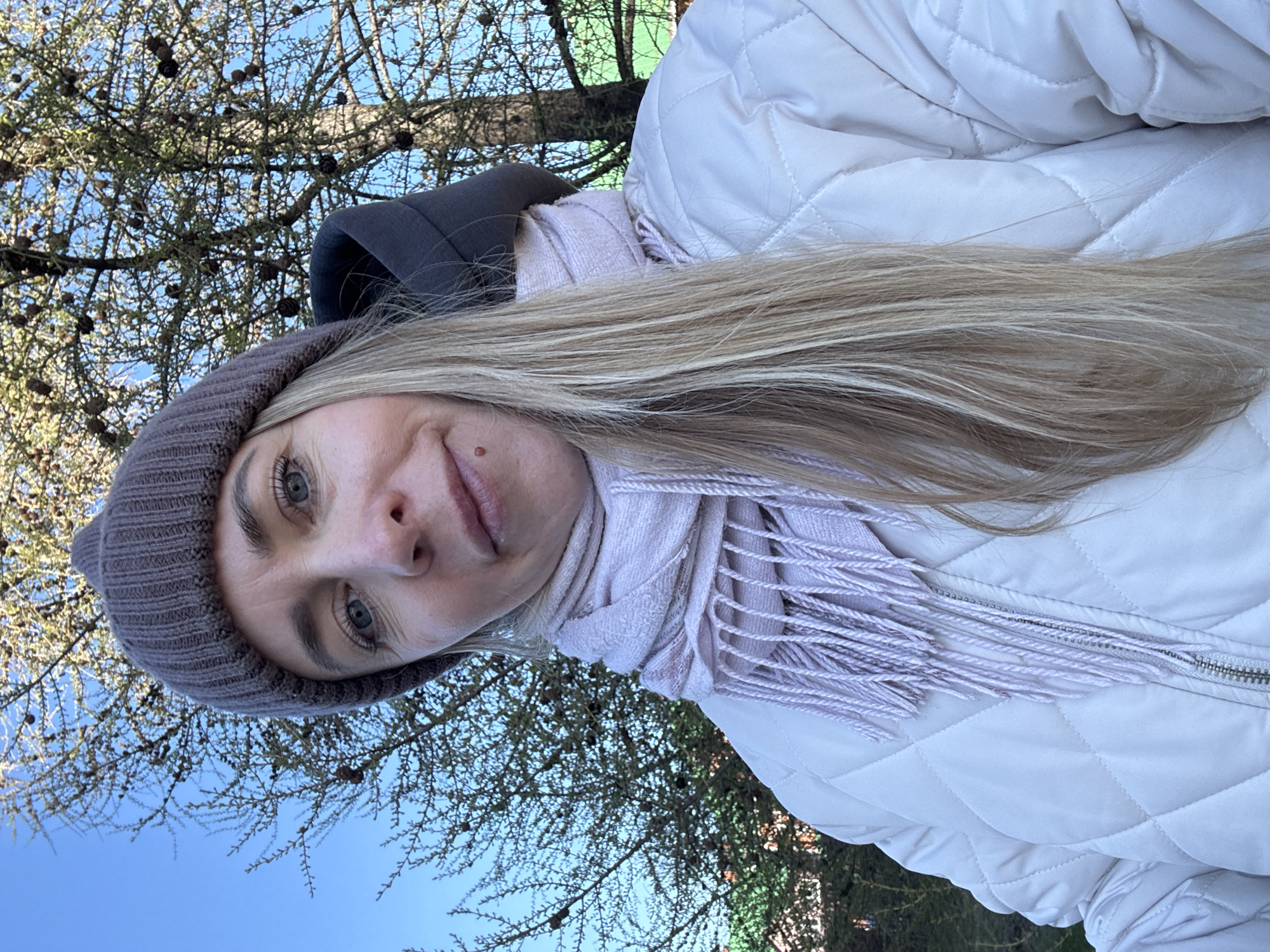 The image shows a selfie of a woman with light skin, blue eyes, and long blonde hair. She is wearing a gray beanie, a light purple scarf, a white quilted jacket, and a dark gray hoodie underneath. The background reveals tree branches with small cones against a clear blue sky. Based on the presence of cones on the branches, the trees are likely coniferous. Given the request for a location within the city of Chaikovsky, Russia, the scene could be located in a park or wooded area within the city, possibly a park like "Park Kultury I Otdykha" or near the Kama River where trees are present.