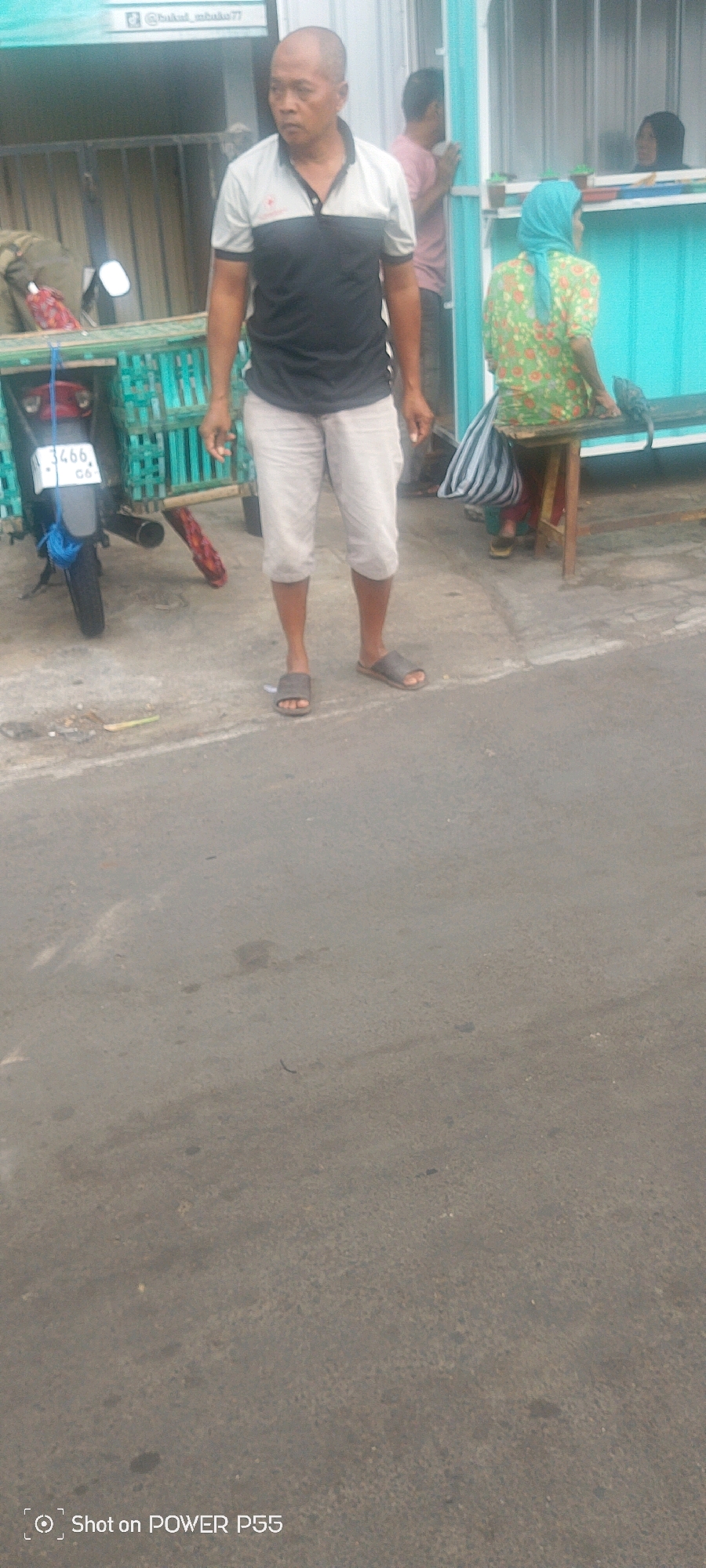A man with a shaved head, wearing a dark gray and white polo shirt and light-colored capri pants, stands on a paved surface. He is looking to his left. He is wearing dark gray flip-flops. To his left, a motorcycle with a license plate reading "3466 CO" is parked next to a teal-colored cart with green mesh sides. In the background, a woman wearing a blue headscarf and a green floral shirt sits on a stool. Behind her, another person in a pink shirt leans against a teal-colored wall, and a woman wearing a black hijab is partially visible through a window. The scene appears to be outdoors in a commercial or street area.