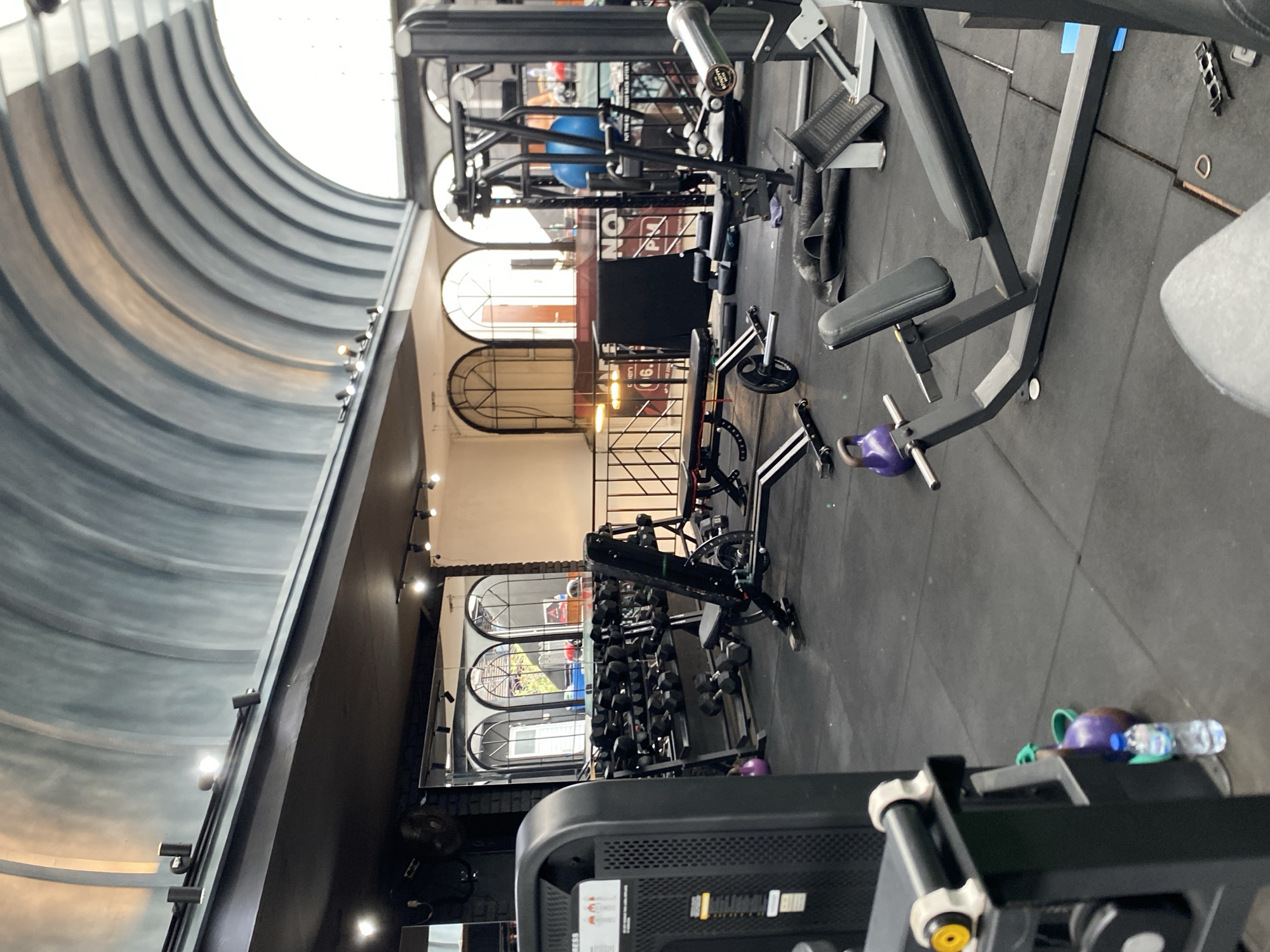 This image depicts the interior of a gym located in North Kuta, Indonesia. The gym features a distinct arched ceiling with a gray and blue striped pattern, illuminated by track lighting. The flooring is black rubber matting, common in fitness facilities.
Prominently featured are various exercise machines and equipment. To the left, a large black weight machine with a display panel is visible. In the center, a rack of dumbbells is neatly arranged, with a weight bench positioned in front of it. Further to the right, another weight machine, possibly a leg press, is present. Kettlebells, one purple and one teal, are also scattered on the floor. A water bottle sits next to the teal kettlebell.
Mirrors line a portion of the wall behind the dumbbell rack, reflecting arched windows with decorative metalwork. Through these windows, greenery and parts of buildings are visible, suggesting an outdoor area beyond the gym. The overall atmosphere is functional and geared towards physical training. No people are present in the image.