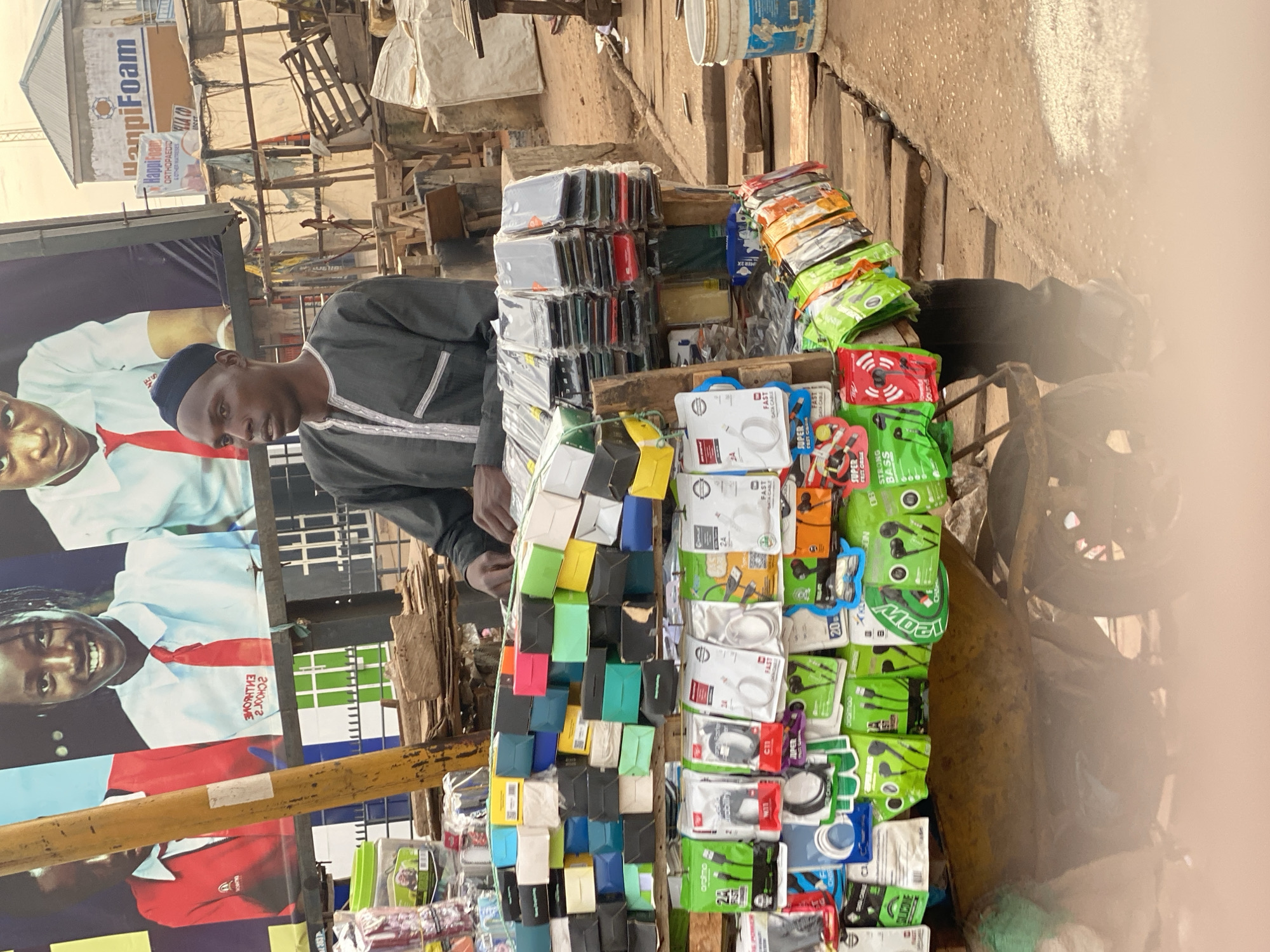 A medium shot captures an outdoor market stall in Sabon Tasha, Nigeria. In the foreground, a man wearing a dark cap and a grey tunic stands behind a wooden stall laden with mobile phone accessories. The stall displays an array of colorful boxes, phone cases, charging cables, and earphones, some in plastic packaging. A weathered yellow pole is visible to the left of the man. In the background, a large advertisement featuring smiling children in school uniforms is mounted on a wall. Further behind, market stalls, wooden crates, and a building with a "HanpiFoam" sign suggest a bustling commercial area. The ground is a mix of dirt and sand, with a partial view of a yellow wheelbarrow and a white bucket in the lower right corner.