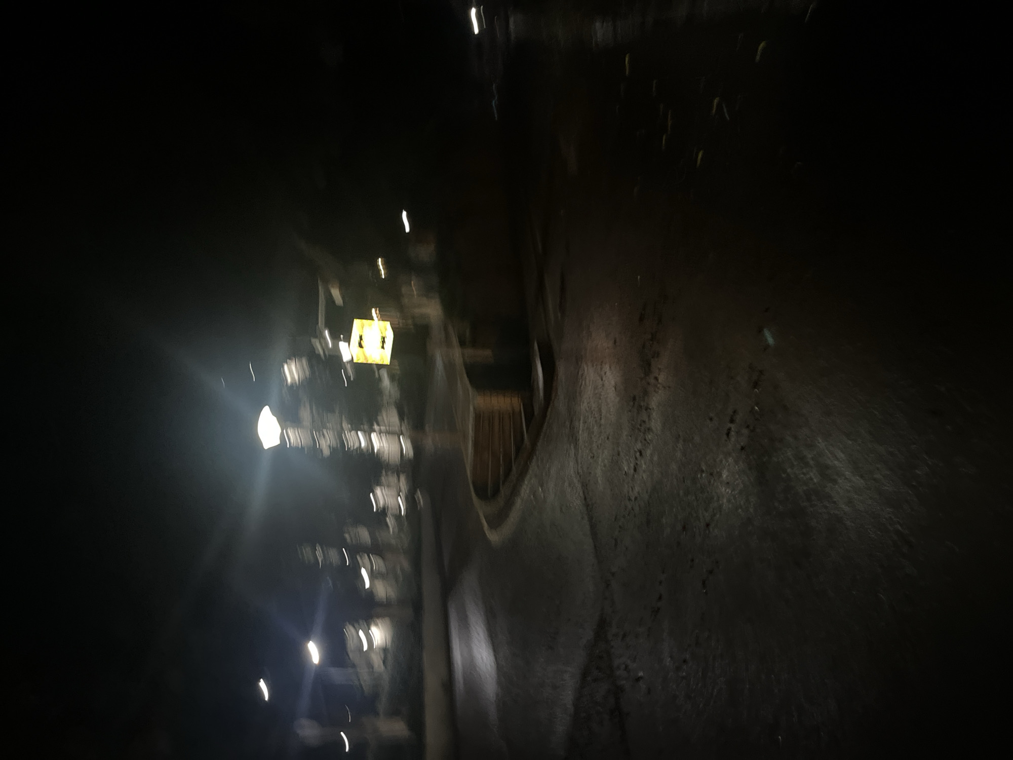 The scene depicts a wet, urban environment at night within Yuzhong District, China. The image is heavily blurred, suggesting motion or low-light conditions with camera shake. Multiple artificial light sources, consistent with streetlights, appear as bright, elongated white streaks and flares across the upper and middle sections of the frame, illuminating portions of the scene. A prominent, intensely bright light source is visible in the upper-right quadrant. In the middle-right, a distinct yellow, square-shaped object, likely an illuminated sign, is visible but also blurred.
The lower half of the image features a dark, reflective paved surface, indicative of a wet road or pathway, with visible puddles and textures. This surface curves from the center-left towards the right, leading into deeper darkness. Along the right edge of this paved area, a structured element consisting of multiple tiers or steps, possibly made of wood or dark stone, is partially visible and curves with the path. The background is indistinct, showing heavily blurred vertical shapes consistent with trees or buildings, largely obscured by darkness and blur. No people, vehicles, or specific actions or interactions are discernible in the frame.