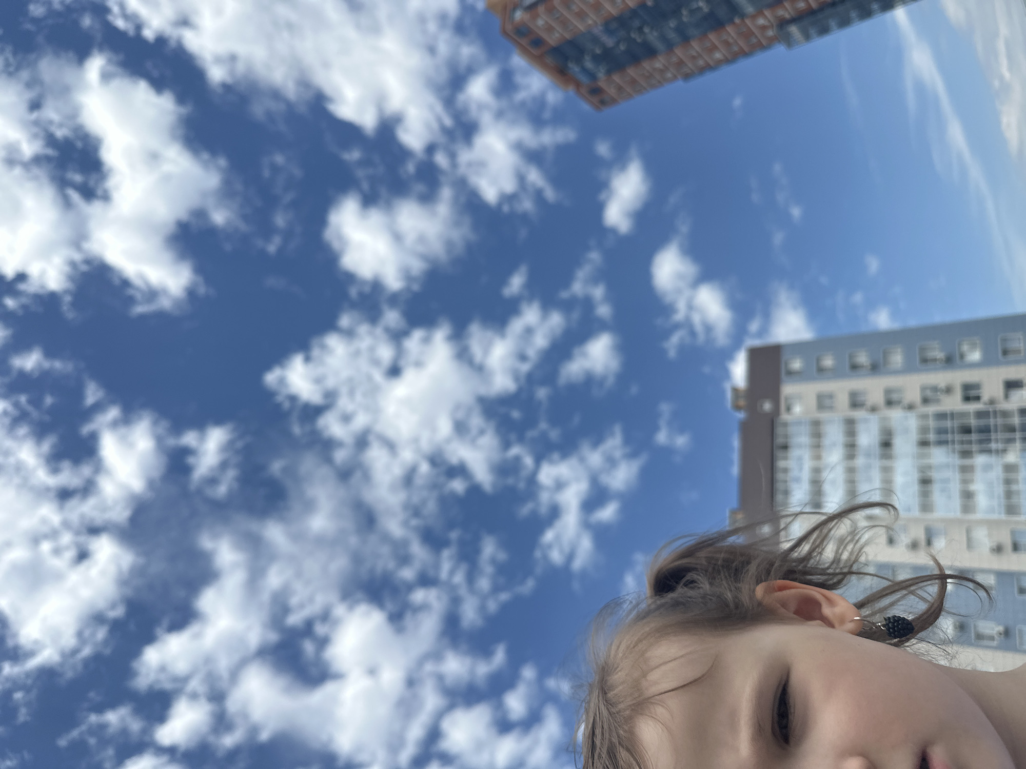 The image shows a low-angle view of a young child outdoors, with a blue sky dotted with white cumulus clouds forming the background. Two buildings are partially visible in the upper part of the frame. One has a reddish-brown facade with dark windows, while the other has a gray and white facade with numerous windows. The child's face is positioned in the lower-right corner of the image, partially obscuring the view. The child has fair skin, light brown hair pulled back in a ponytail, and is wearing a dark earring. The child is positioned between apartment buildings with white windows and blue sky and white clouds. Based on architectural style, location and context clues, the image was likely captured in Khabarovsk, Russia.