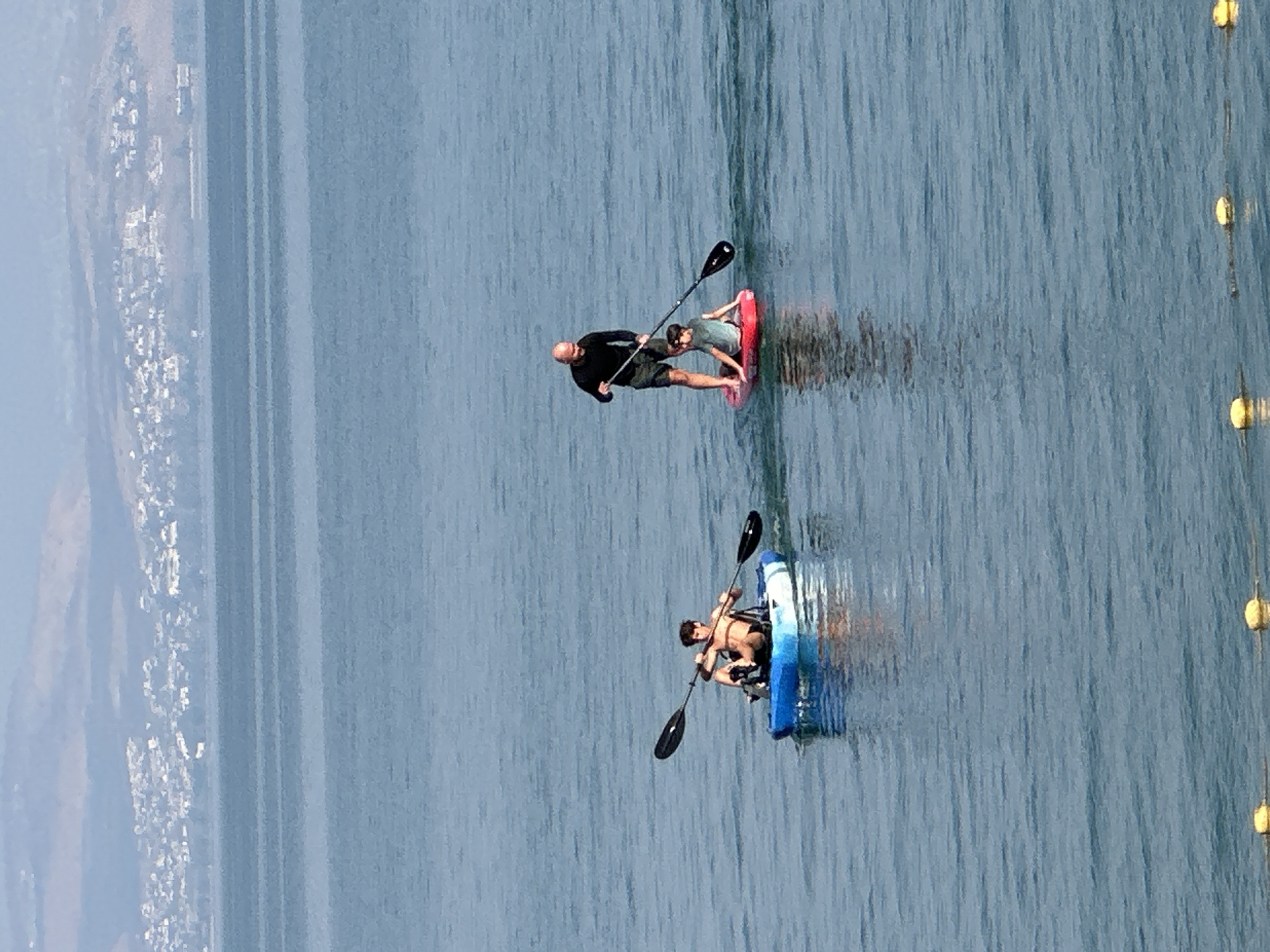 The image depicts a wide shot of the Sea of Galilee near Tiberias, Israel. In the foreground, two individuals are engaged in water activities. On the left, a young man is in a blue and white kayak, paddling with a double-bladed oar. He is wearing dark shorts. On the right, an adult man is standing on a red paddleboard, holding a paddle. A child is sitting on the front of the paddleboard. The man is wearing a black long-sleeved shirt and shorts. In the background, the city of Tiberias is visible as a cluster of white buildings along the coastline, nestled against rolling hills. The water is a calm, muted blue, with subtle ripples. The sky is hazy and light.