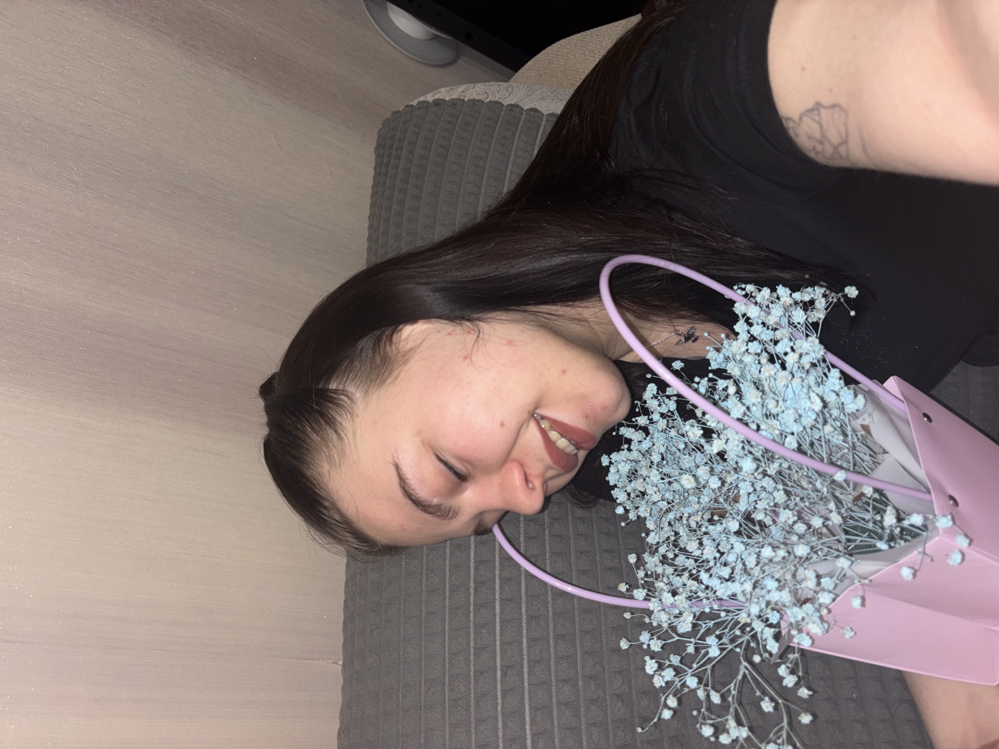 A woman is lying on a gray textured surface, possibly a couch or bed, with her head resting near the left edge of the frame. She has dark hair and is wearing a black shirt. Her face is turned to the side with a slight smile. She holds a small pink shopping bag containing light blue baby's breath flowers. The background includes a neutral-colored wall and a partially visible monitor in the upper portion. There is no indication in the photo that the location is Perm, Russia.