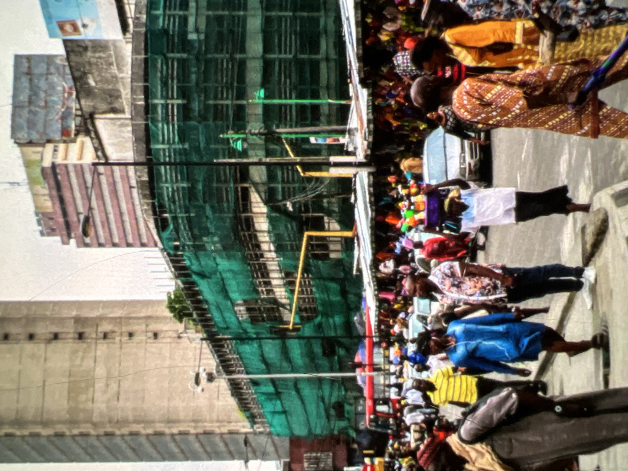 This is a daytime, eye-level, outdoor medium shot of a busy street scene in Lagos, Nigeria. The foreground features several individuals walking on a paved street. To the left, a person wearing a brown coat and a tan scarf walks away from the camera. In the center, a woman in a bright blue dress and white earphones walks towards the right. To her right, a man in a floral-patterned shirt and dark pants walks towards the right. Further back, a man in a yellow striped shirt walks towards the left. In the background, there is a market with stalls covered by striped awnings. Various people are visible browsing the stalls. A red bus is partially visible behind the market stalls. In the far background, there are several multi-story buildings, one of which is covered in green scaffolding. The sky is overcast. The overall impression is one of a vibrant and active urban environment.