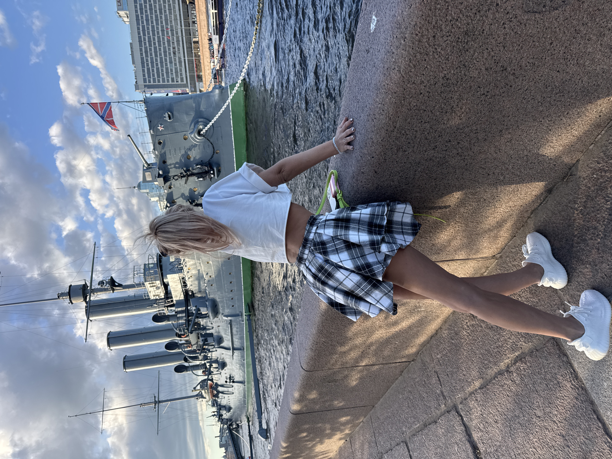 In Saint Petersburg, Russia, a blonde-haired woman with her back to the camera leans on a granite embankment, looking towards the Aurora cruiser. She wears a white crop top and a plaid skirt. The cruiser, a museum ship and historical landmark, is moored on the Neva River, with several other ships visible in the background. The sky is partly cloudy, and the water is choppy. The scene is captured on a sunny day, with shadows cast across the embankment.