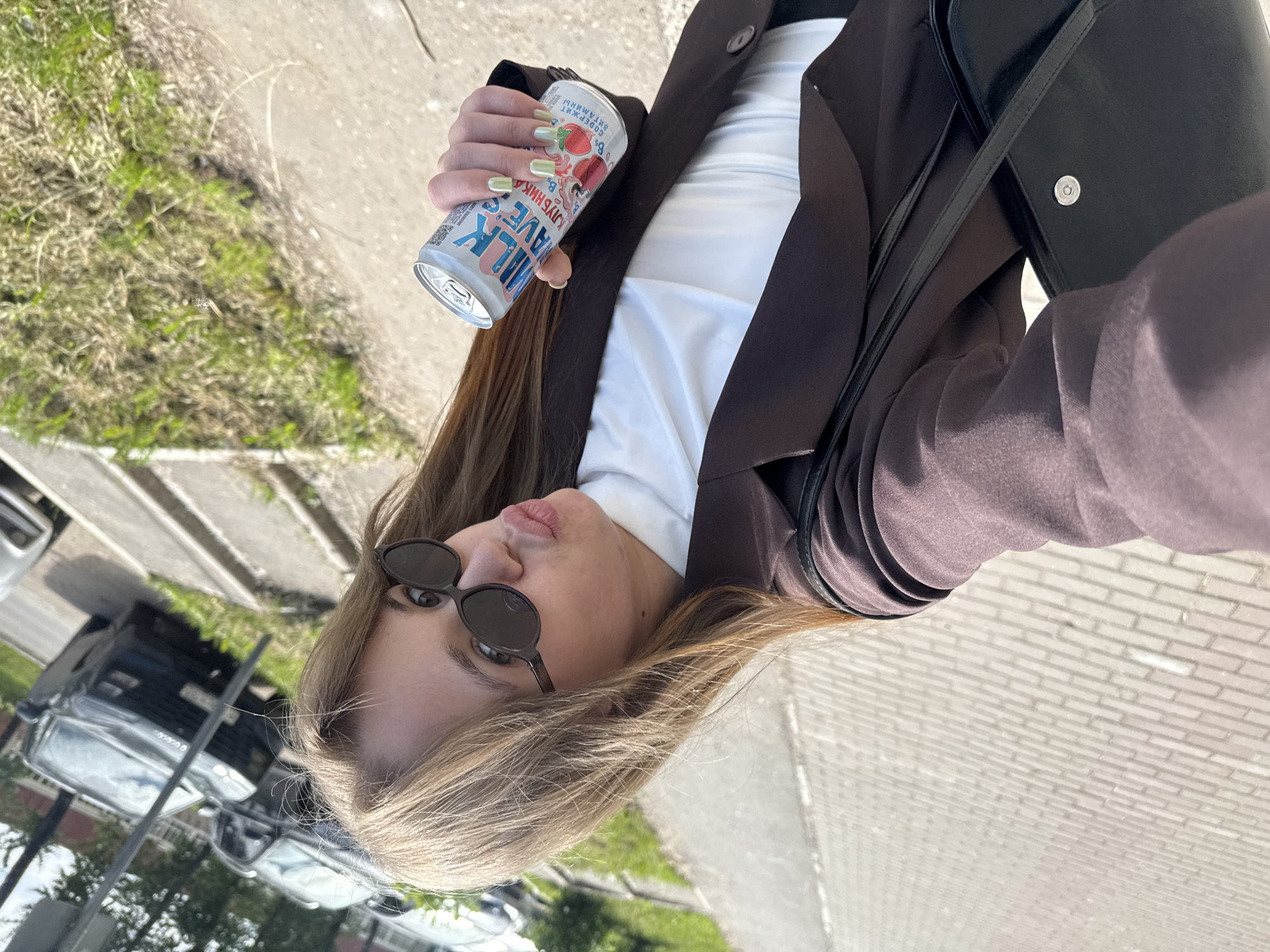 The scene is set outdoors in the city of Chaikovsky, Russia, depicting a bright daytime environment. A young woman, positioned centrally and angled diagonally, is visible from the chest up, holding a beverage can and striking a pose for a selfie. She has light brown hair, wears dark round sunglasses, and is making a puckered lip expression towards the camera. Her right hand, adorned with light green nail polish, firmly grasps a light-colored metal can. The can's label displays "MILK WAVE" and "КЛУБНИКА" (Strawberry) in Russian text, alongside an image of strawberries and other small Russian text. She is dressed in a white top beneath a dark brown/maroon blazer, with a black shoulder bag visible over her left shoulder. The ground beneath her is a paved sidewalk constructed from light-colored rectangular bricks. Beyond the sidewalk, to the right, is a strip of grass, and further back, a street with several parked vehicles, including dark-colored sedans and a partial view of a white car. Sparse trees line the background. The lighting suggests a clear, sunny day.