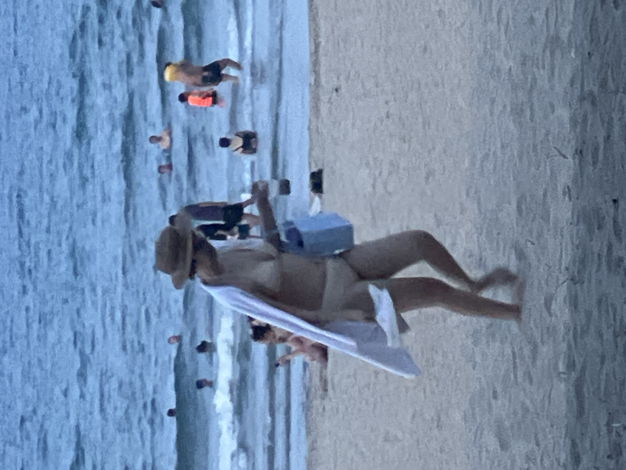 The image depicts a beach scene in Đà Nẵng, Vietnam, with the ocean in the background and sand in the foreground. A woman in a bikini and a straw hat is walking on the sand, carrying a blue bag and a white towel draped over her shoulder. Several other people are visible in the shallow water, some swimming and others standing. The water is calm with small waves. The overall lighting suggests it is either early morning or late afternoon.