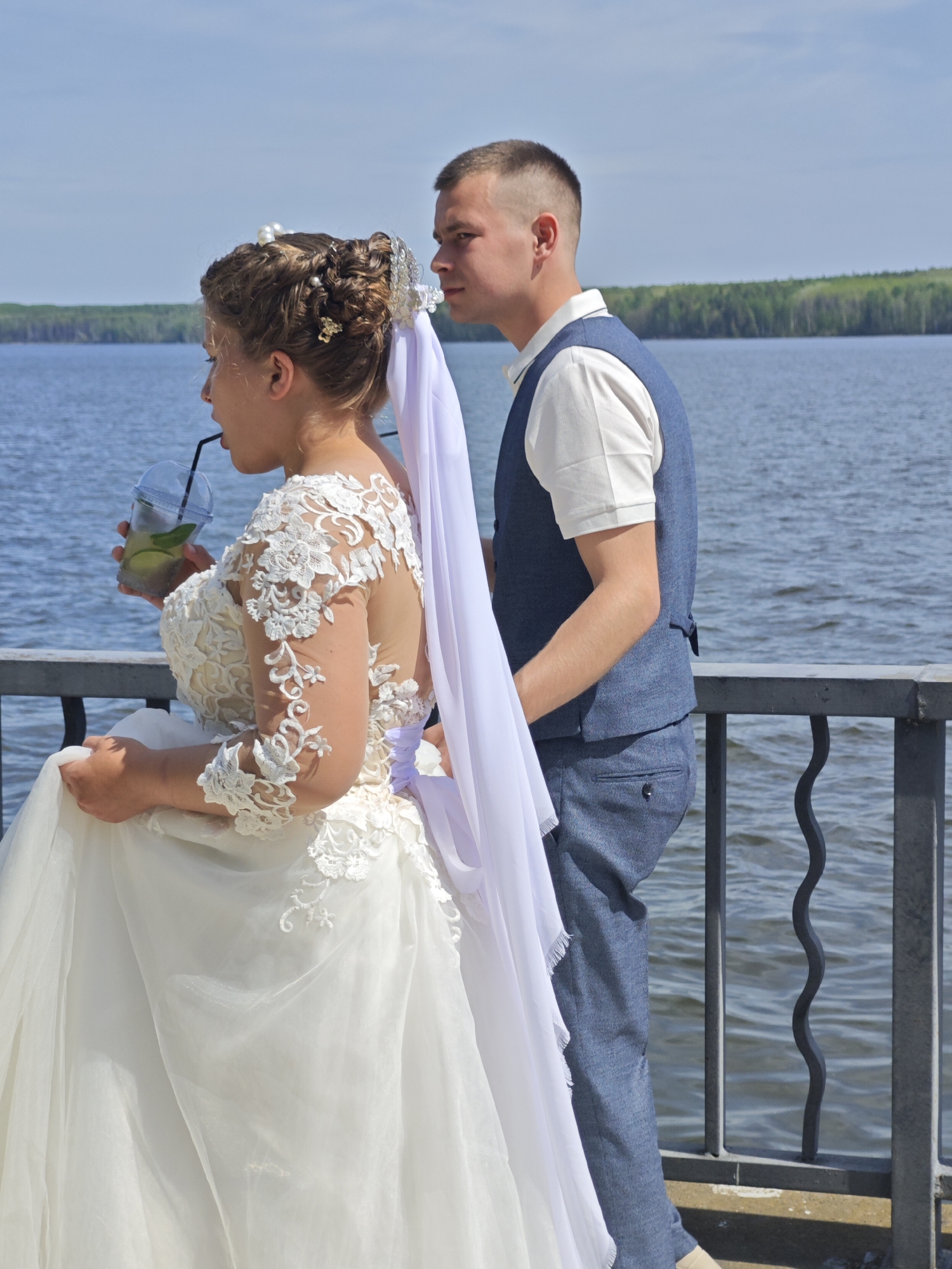A bride and groom stand next to a grey metal railing overlooking a body of water, likely the Kama River. The bride, on the left, wears a white lace wedding dress with a long white veil. She is drinking from a clear plastic cup with a straw, containing a drink with lime slices. The groom, on the right, wears a light blue suit with a white polo shirt. The background features water with light ripples and a distant green shoreline with trees under a bright, clear sky, suggesting it is daytime. The location is likely in Chaikovsky, Russia.