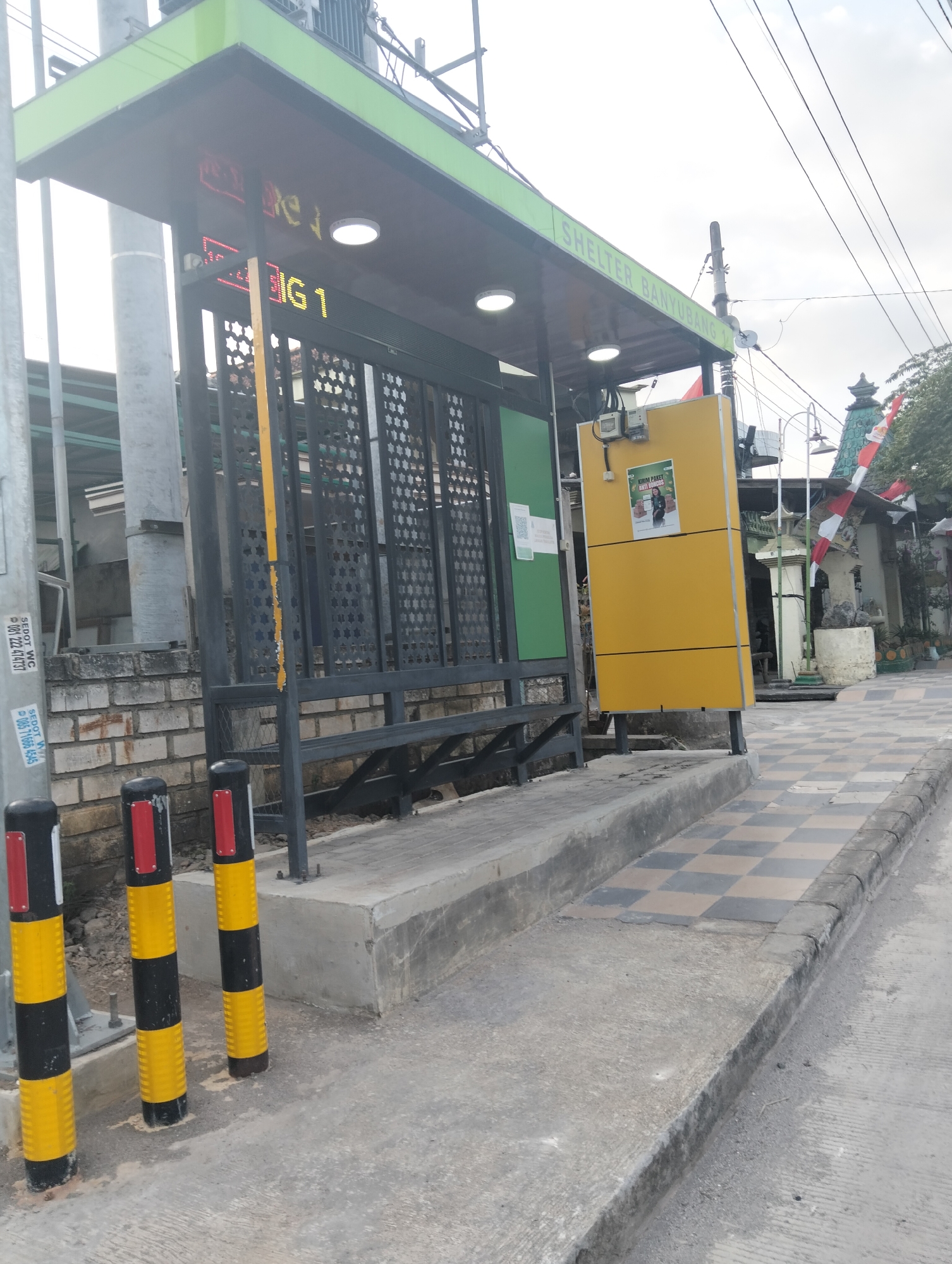 The image shows a bus shelter in Dagan, Indonesia, with the words "SHELTER BANYUBANG 1" visible on the side. The shelter has a green roof and a dark-colored frame with a black metal grate. A yellow cabinet is attached to the side of the shelter, displaying a poster with a woman's image and text in Indonesian. Three bollards, painted black and yellow with red reflective strips, are positioned in front of the shelter. The ground in front of the shelter is paved with concrete and a checkered pattern of light and dark tiles. In the background, there is a brick wall, some greenery, and Indonesian flags. The sky is overcast.