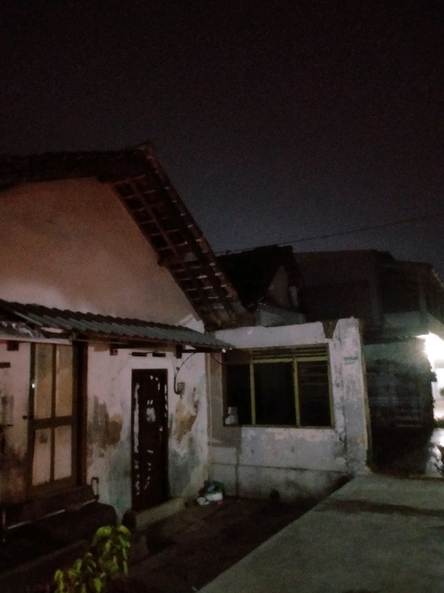 The image depicts a nighttime view of a modest building in the city of Kasihan, Indonesia. The structure appears to be a house or a small commercial establishment with a sloped roof covered in what seem to be clay tiles. The walls are weathered, showing signs of age and possibly some damage.
In the foreground, a section of a dark-colored bench is visible, positioned in front of a doorway with a dark-colored door. To the left of the door, there is a window with a yellow frame. The ground in front of the building is a concrete or asphalt surface, with some scattered debris, including what appears to be a plastic bag and possibly some footwear on the right side. A small plant with green leaves is growing in the lower-left corner of the image.
The lighting suggests it is dark, with a faint ambient light likely coming from the sky or distant artificial sources. The overall impression is that of a simple, lived-in dwelling or business in a residential or mixed-use area. There are no people visible in the frame. The architecture and materials are consistent with common construction styles found in many parts of Indonesia.