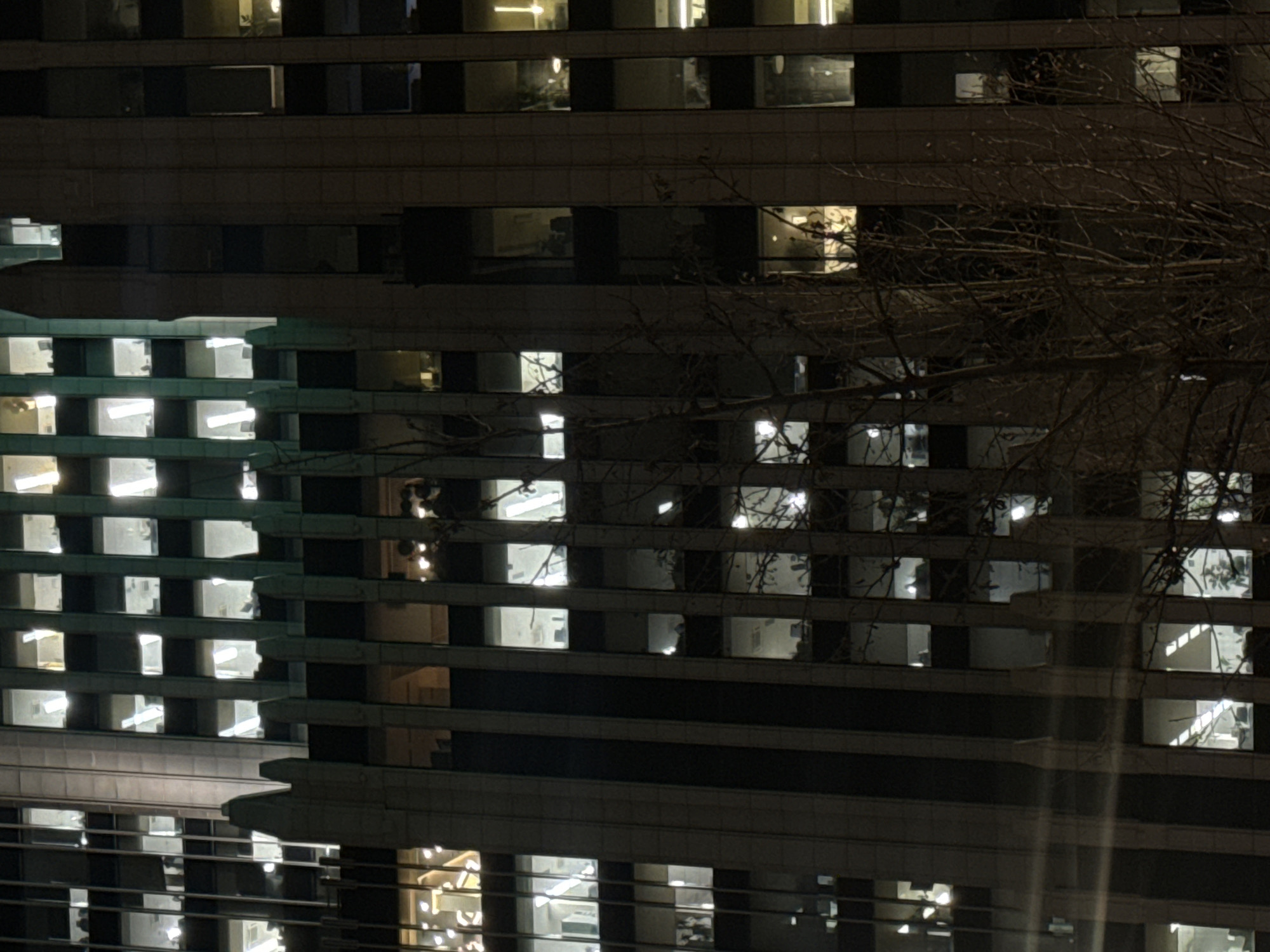 The image displays a nighttime, vertical view of the former Ministry of Aviation Industry building, located at Ulitsa Kuznetsky Most, 3/2с1, Moscow, Russia. The multi-story structure is illuminated by interior lights, revealing distinct architectural sections. The central lower-to-mid portion of the building features a lighter-toned, possibly stone-clad facade with vertical pilasters and a crenelated parapet. Above this, and to the right, a significant section of the building exhibits a dark green or teal-hued facade with a regular grid of square windows. To the far left, a separate, more modern-looking section of a building with a dark, grid-patterned glass facade and rectangular windows is visible.
Numerous windows across all visible sections of the building are illuminated, casting white or warm yellow light. Within these illuminated windows, faint details of office interiors are discernible, including linear fluorescent light fixtures, desks, chairs, and computer monitors. No individuals or specific actions are clearly identifiable within the windows. Partially obscuring the building in the lower-mid and right foreground is a bare, deciduous tree with an intricate network of branches, silhouetted against the illuminated facade. The environment is an urban setting at night.