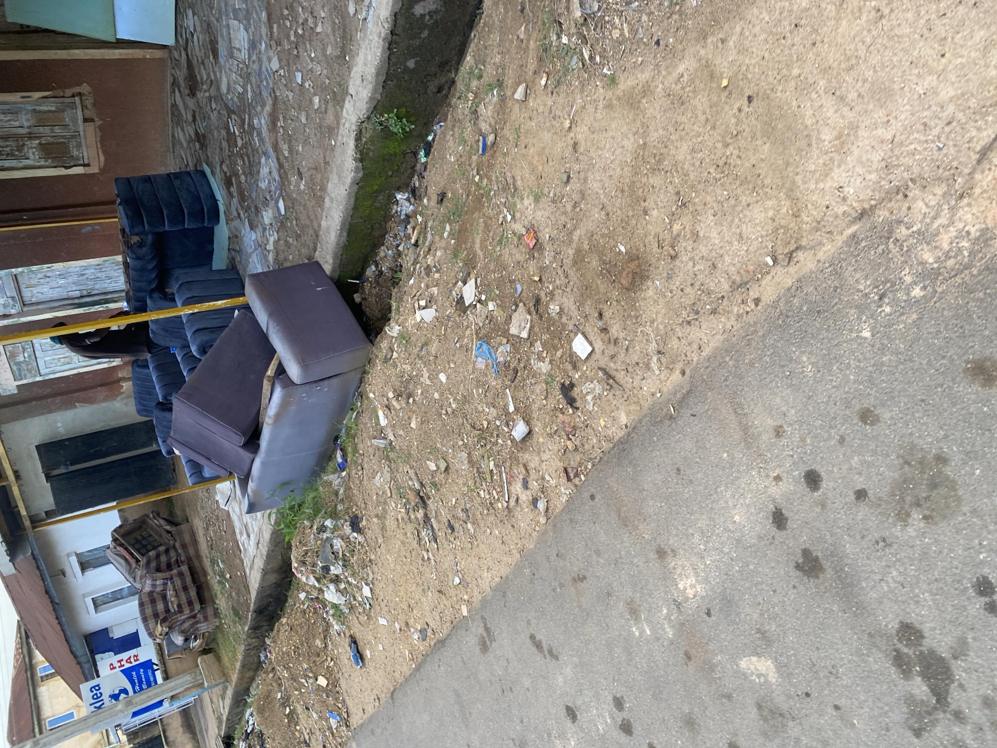 The image shows a street scene in Jos, Nigeria. Several dark blue upholstered sofa sections are stacked on the left side of the frame, some appearing to be damaged or discarded. Adjacent to the sofas, on the right, is a dirt embankment cluttered with various pieces of debris, including plastic bags, paper, and other unidentifiable trash, extending down to a narrow, concrete-lined drainage channel. A paved road runs along the bottom right of the image, with scattered dark stains.
In the background, a building with brown walls and barred windows is visible. A red-tiled roof extends from the building on the left. A partially visible sign with blue lettering reads "kIea" and another smaller sign below it shows "PHAR" in blue letters on a white background, with "Health & Beauty" and a phone number below that. A striped blanket is draped over what appears to be another piece of furniture or debris in front of the building.
The scene appears to be an outdoor setting, likely a street or roadside, given the presence of the road and the buildings. The lighting suggests it could be daytime, with no strong shadows indicating a specific time. The presence of debris suggests a potentially neglected or underserved area. No people are visible in the frame.