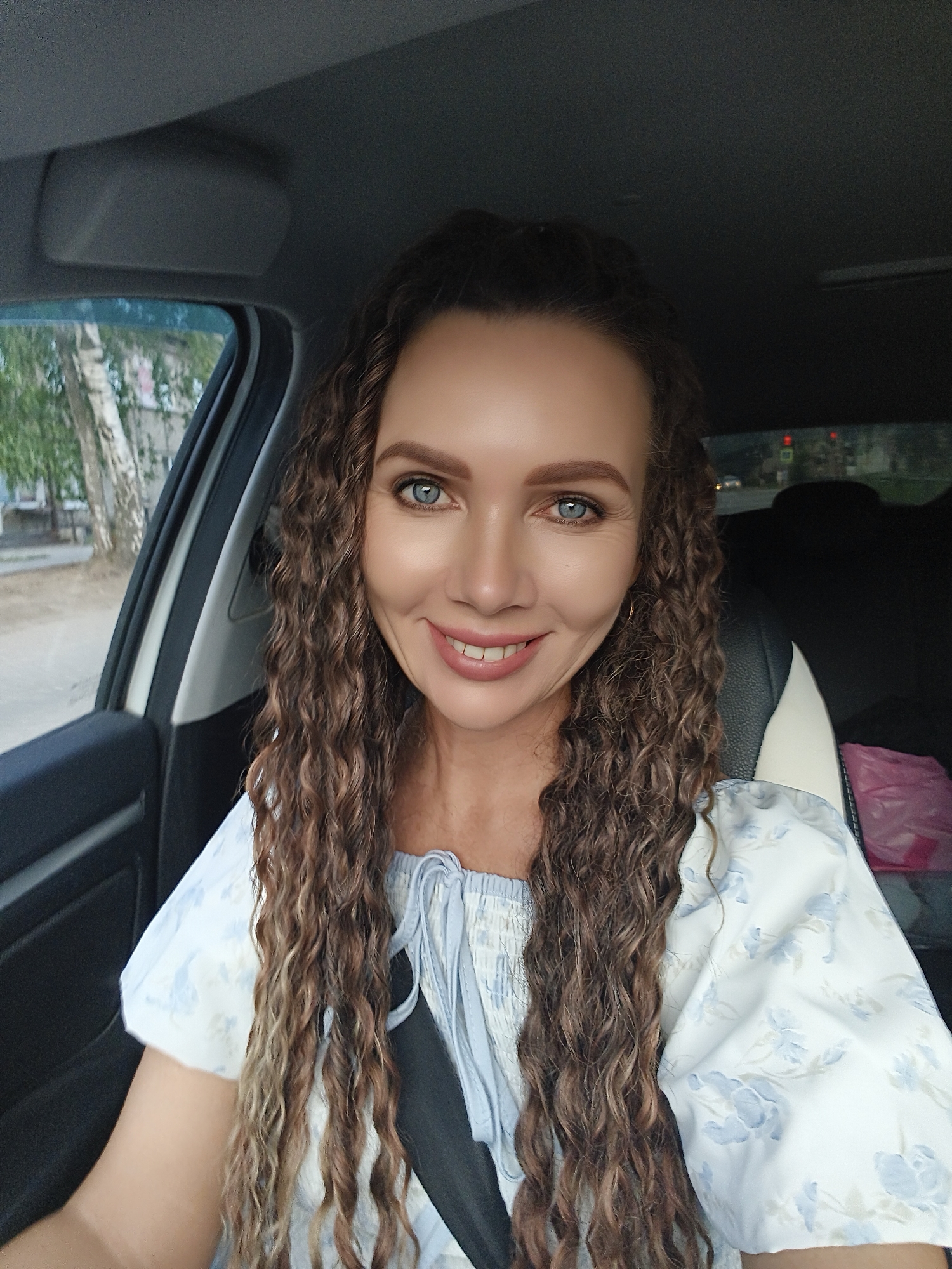 A woman with long, curly brown hair and blue eyes is sitting in the driver's seat of a car. She is smiling and looking at the camera. She is wearing a white and blue floral print shirt. The car appears to be parked on a street in the city of Chaikovsky, Russia. Trees and buildings are visible outside the car window. The interior of the car is dark, with a black and white seat and a grey headliner.