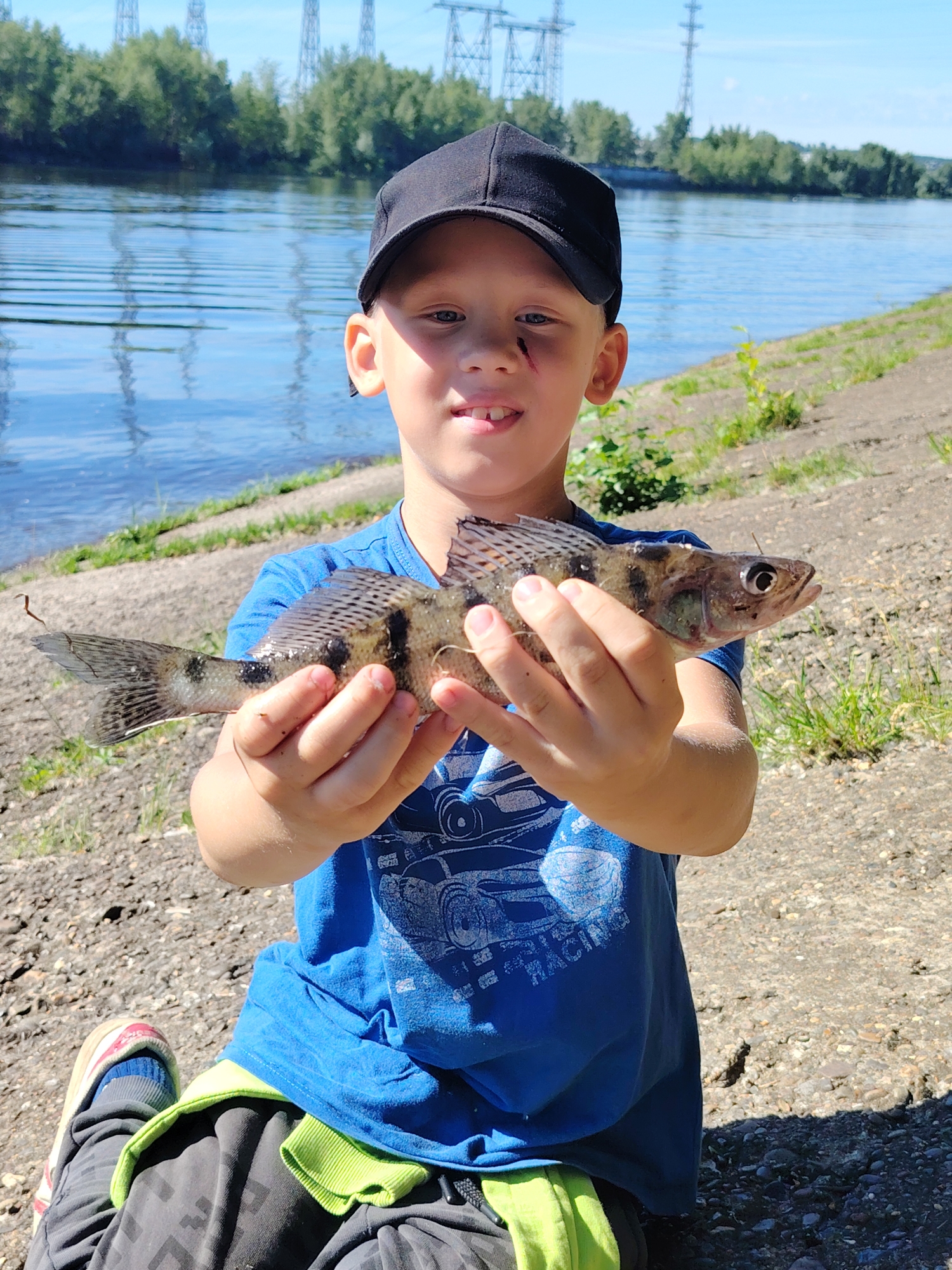A young boy wearing a black baseball cap and a blue t-shirt sits on the rocky bank of a river, holding a small fish he has caught. The boy is smiling, showing his teeth, and has a small scrape on his cheek. He is dressed in dark pants with a bright green waistband and colorful athletic shoes. The t-shirt has a graphic print on it, featuring a car and the word "RACING".
The background shows a wide river with gentle ripples reflecting the clear blue sky. The opposite bank is lined with lush green trees. In the distance, large power line towers are visible against the horizon, indicating an industrial or developed area nearby. The sun is bright, suggesting it is daytime, and the overall atmosphere is cheerful and suggests a successful fishing trip. The setting appears to be a natural area on the outskirts of a city, given the presence of both the river and the power structures. The image conveys a sense of childhood accomplishment and outdoor recreation.