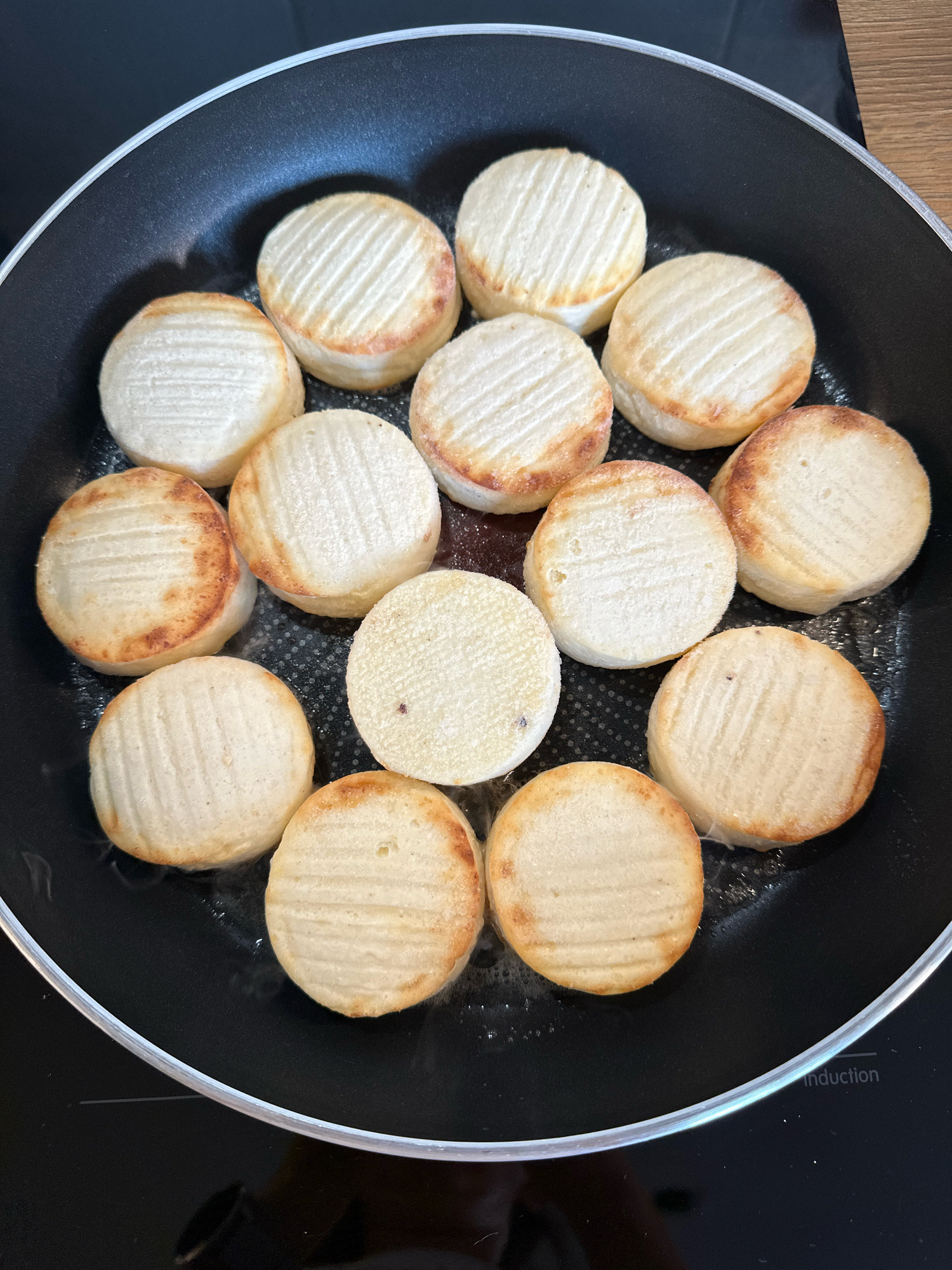 A top-down, close-up view of a non-stick frying pan filled with round, flat slices of what appears to be cheese, possibly halloumi, being pan-fried. The cheese slices are a light beige color with grill marks or ridged texture on their surfaces and are lightly browned on the edges. Some slices have a slightly darker golden-brown hue, indicating they are cooking. The pan is black with a silver rim and is placed on a black induction stovetop, indicated by the word "Induction" visible near the bottom right edge of the image. The overall environment suggests a kitchen setting. The image does not contain any people or any recognizable landmarks or features that would place it in the city of Vidnoye, Russia.