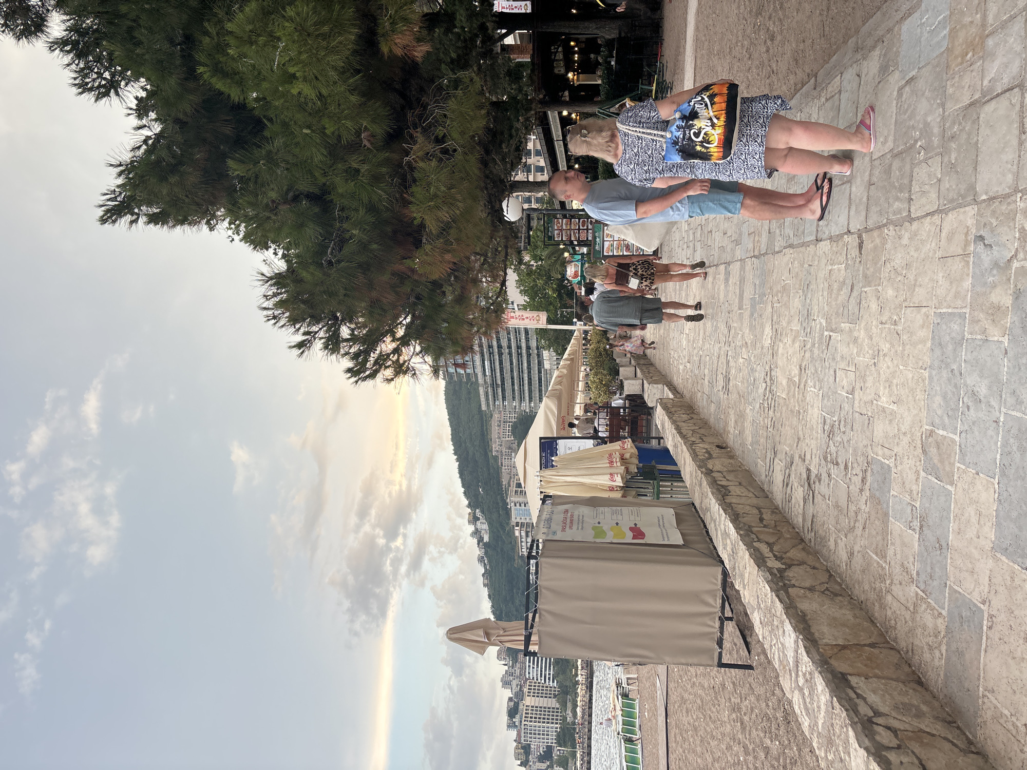 In Budva, Montenegro, a paved walkway runs alongside a sandy beach. On the right, a cluster of lush green pine trees casts shadows over the path. Several people are walking on the walkway. In the foreground, a woman with blonde hair pulled back in a ponytail is wearing a blue and white patterned dress and carrying a tote bag with the words "Summer Travel" printed on it. Beside her, a man in a light blue t-shirt and shorts walks ahead. Further down the path, a couple is walking away from the camera, the woman in a leopard-print skirt and the man in a grey t-shirt and shorts. On the left, a beige tent-like structure stands near the beach, with umbrellas and signs visible. The sea is visible behind the tent, and beyond that, buildings and a hillside covered in more structures rise up in the background under a cloudy sky.