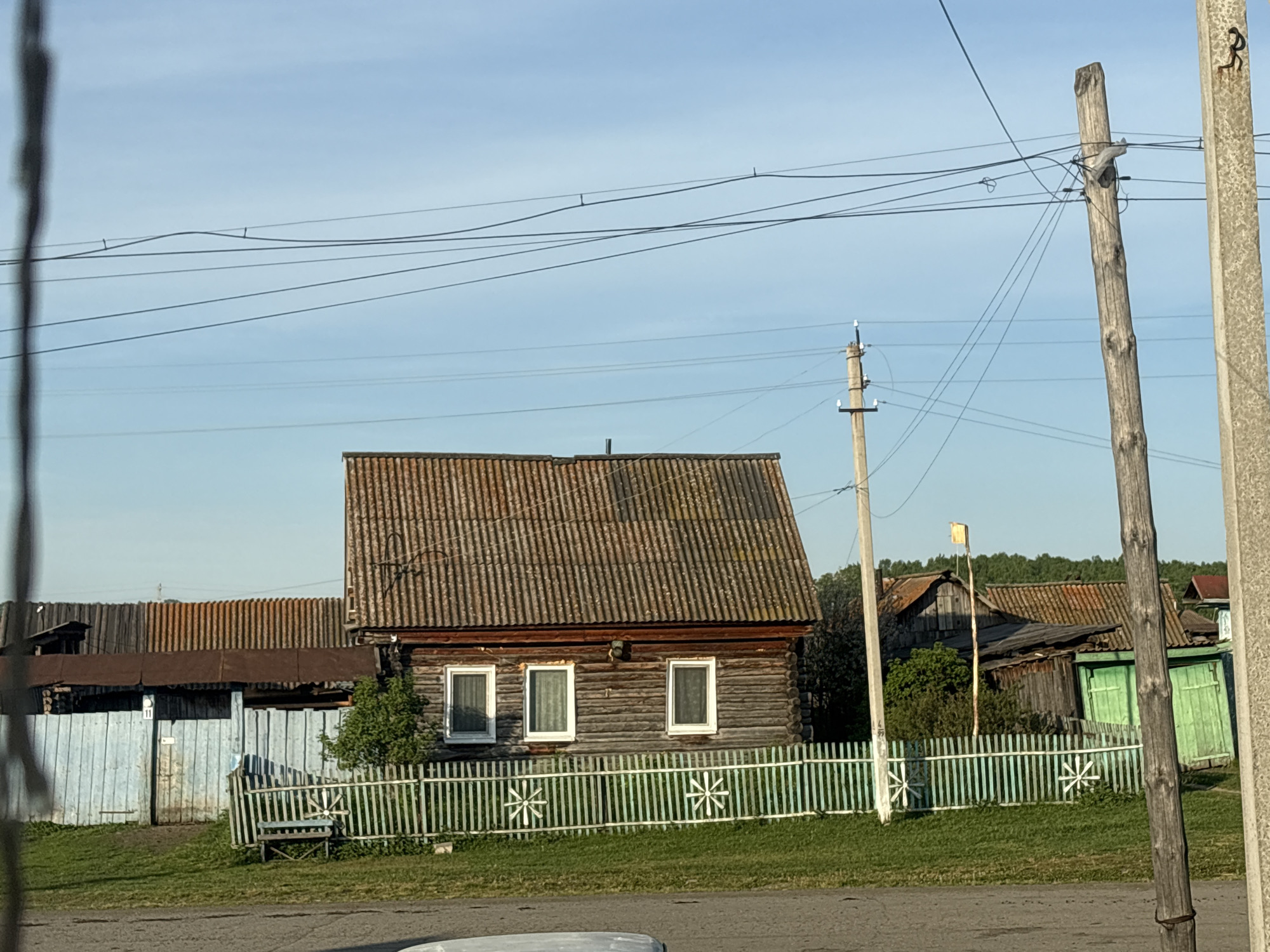 The image shows a rural scene in an unspecified location. The main subject is a wooden house with a brown corrugated roof. The house has three white-framed windows on the front. A pale green and white picket fence surrounds the house. In the background, other houses with wooden structures and corrugated roofs are visible. Utility poles with wires run across the scene, typical of a rural setting. The sky is clear and blue. The overall impression is of a quiet, rustic village.