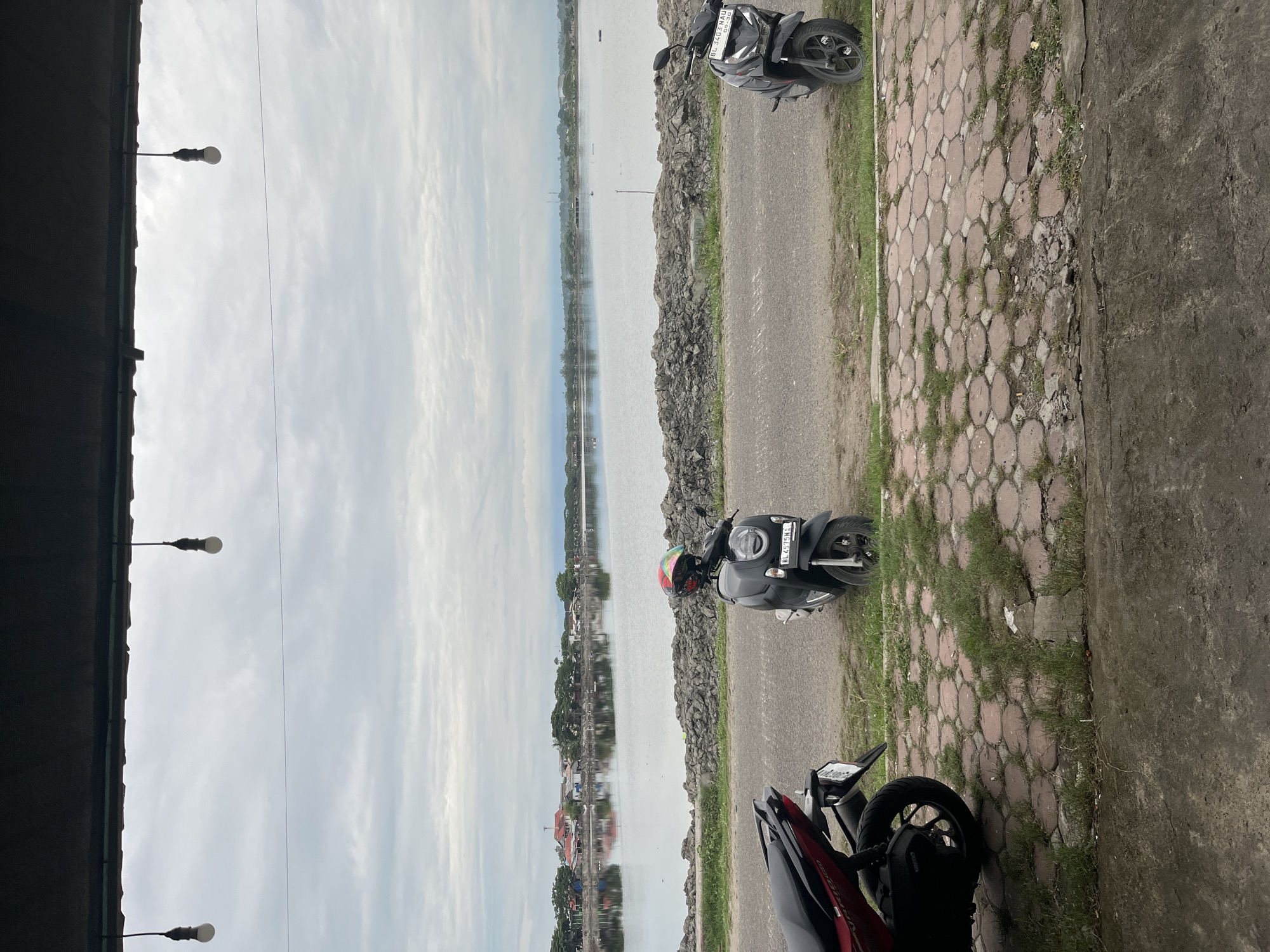The image shows a street scene in Lhokseumawe, Indonesia. A body of water, likely a lake or bay, is visible on the left side of the frame. The street runs parallel to the water, separated by a rocky barrier. There are three motorbikes parked on the side of the road. The sky is overcast. There are buildings on the other side of the body of water. The photo is taken from under a partial structure with streetlights attached.