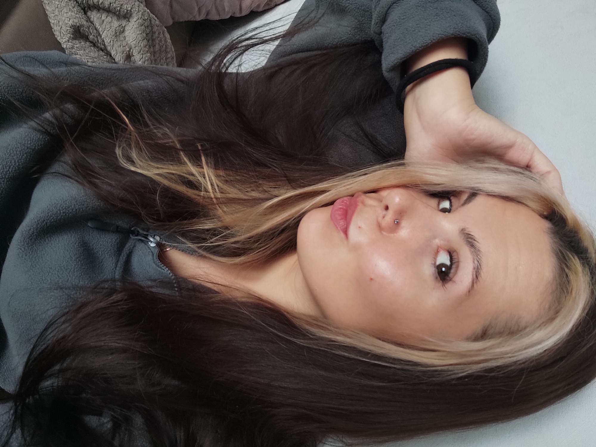 A young woman is lying down, her head resting on her hand. She has long, dark brown hair with a blonde streak at the front. She is wearing a gray fleece jacket and has a black hair tie around her wrist. She has a nose piercing and is looking directly at the camera with a neutral expression. The background appears to be a light gray surface. It is not possible to ascertain the location as Chaikovsky, Russia, from this image alone.