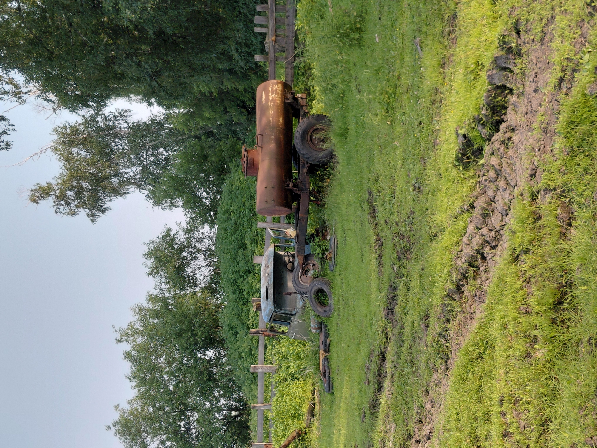 In a grassy, uneven field in Novoselovo, Russia, an abandoned, rusted water tank trailer is hitched to a derelict, light blue truck cab. The truck cab is missing its body and appears to be partially sunk into the muddy ground. Several loose tires are scattered around the truck. A wooden fence runs along the back of the property, with lush green trees and foliage behind it. The sky is clear and pale blue. The scene suggests a rural or semi-abandoned setting, possibly a farm or storage area, where old equipment has been left to decay.