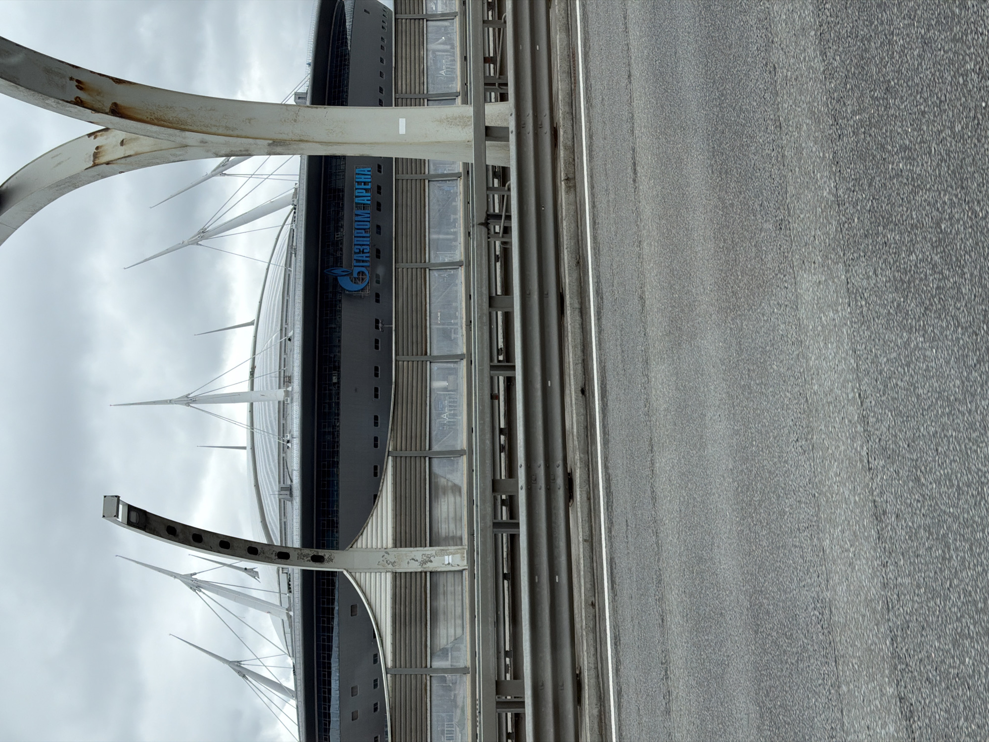 The image shows a view of the Gazprom Arena in Saint Petersburg, Russia, as seen from an elevated road. The arena's exterior is visible, featuring its distinctive architectural design, including the sail-like structures and the Gazprom logo. A guardrail is visible in the foreground, separating the road from the arena. The road surface is gray and appears to be asphalt. The sky is overcast.
